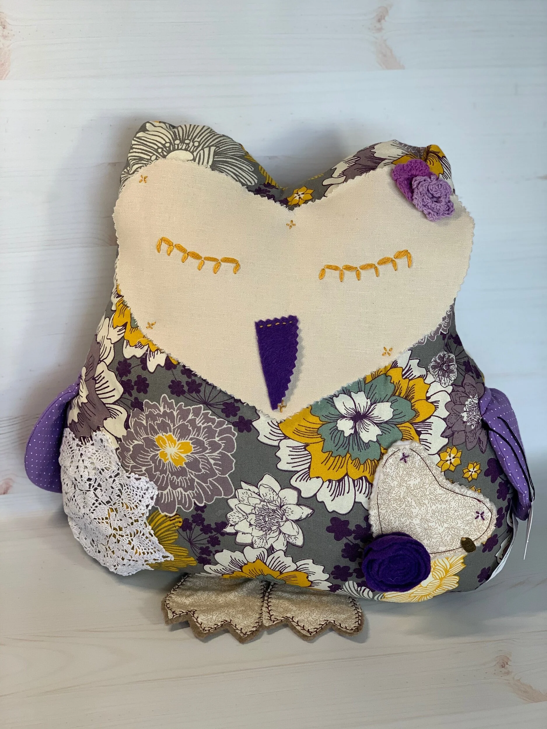 A decorative owl-shaped pillow with purple and yellow floral patterns, featuring embroidered details and fabric flowers, resting on a white wooden surface.