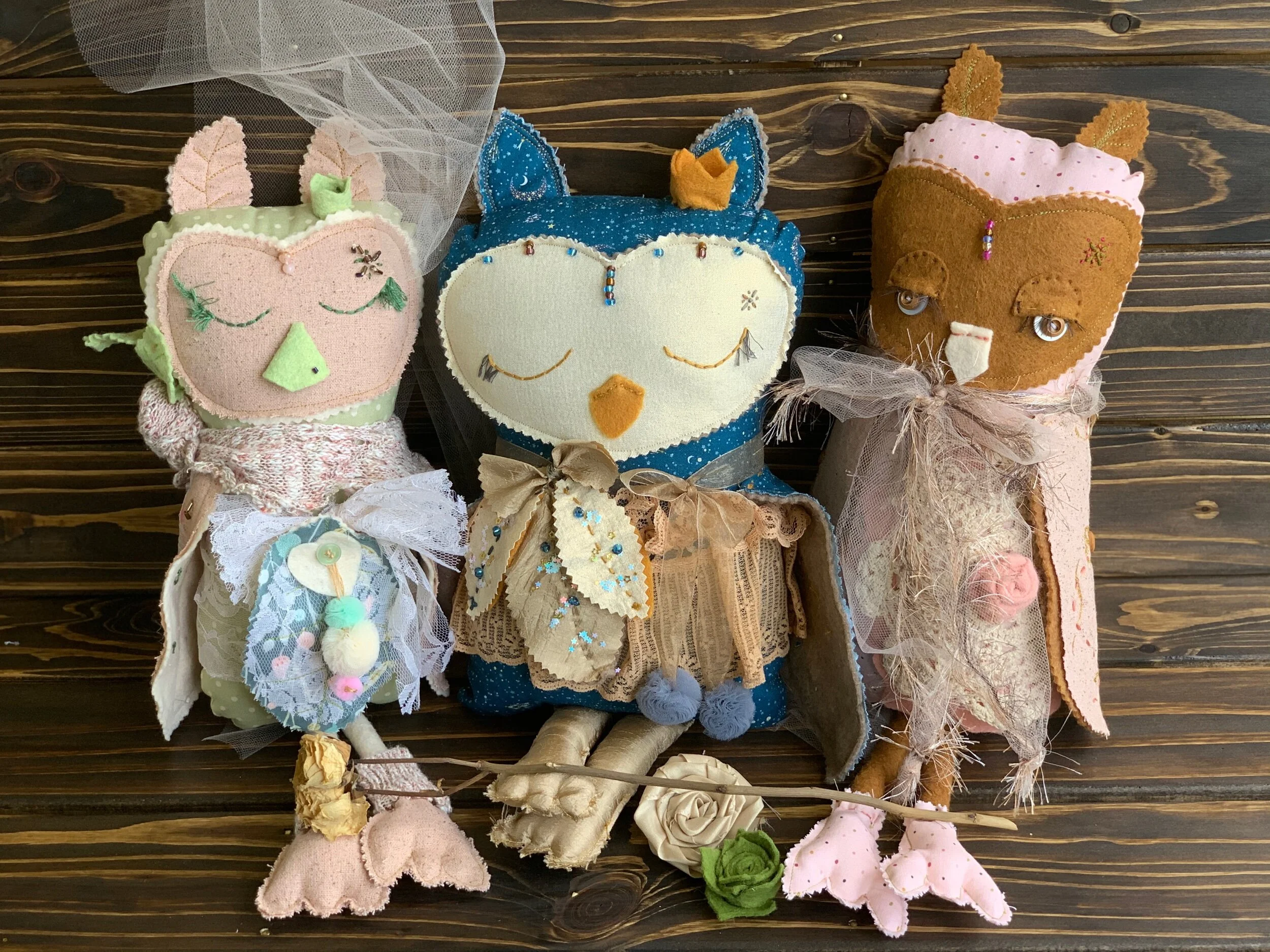 Three handmade plush owl dolls in colorful clothing sitting on a wooden surface.