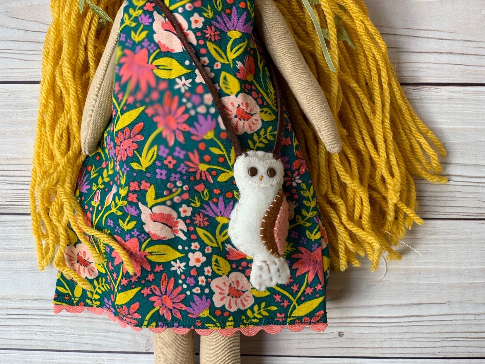 Handmade doll with yellow yarn hair, wearing a colorful floral dress and a small felt owl accessory on a wooden background.