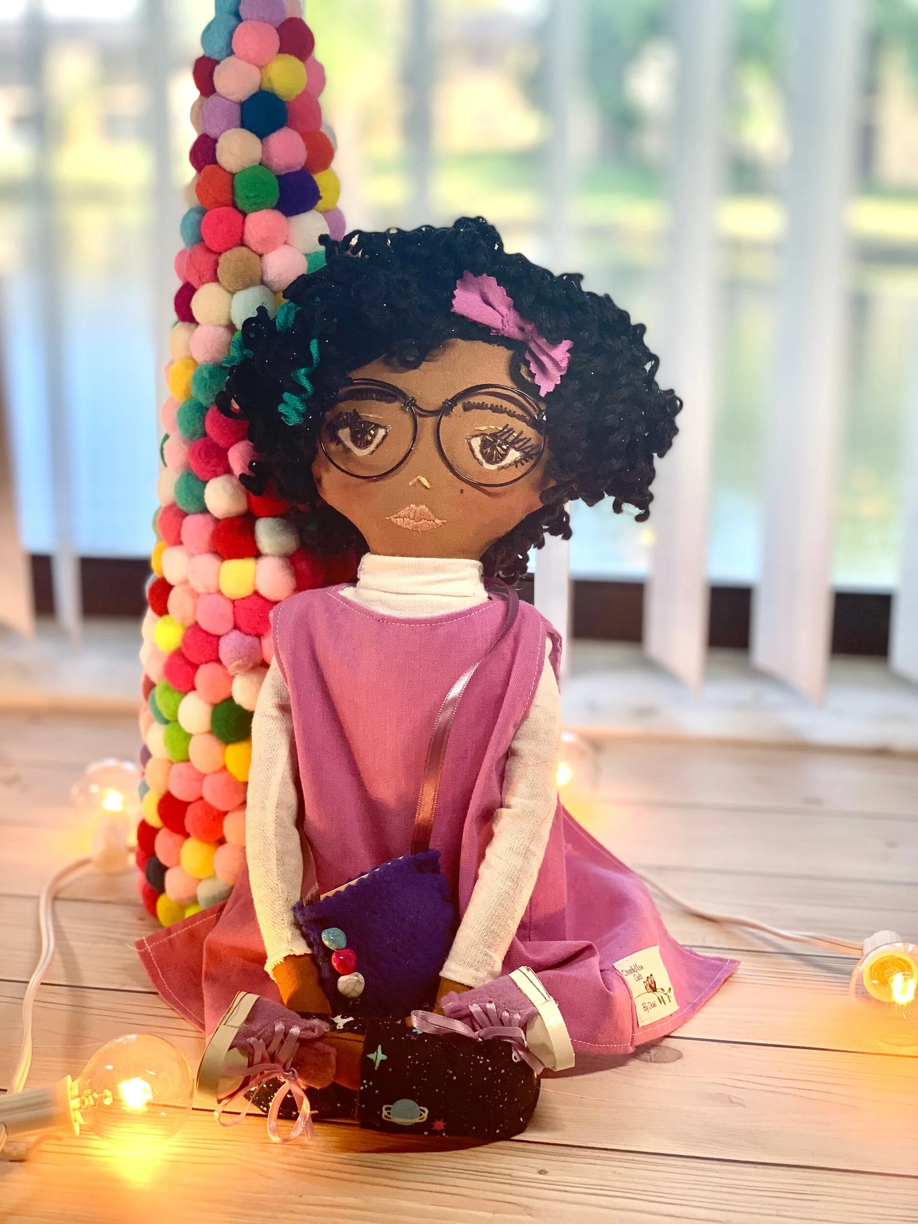 Handmade doll with glasses, curly black hair and a purple dress, seated in front of a colorful pom-pom cone, with decorative lights on wooden floor.