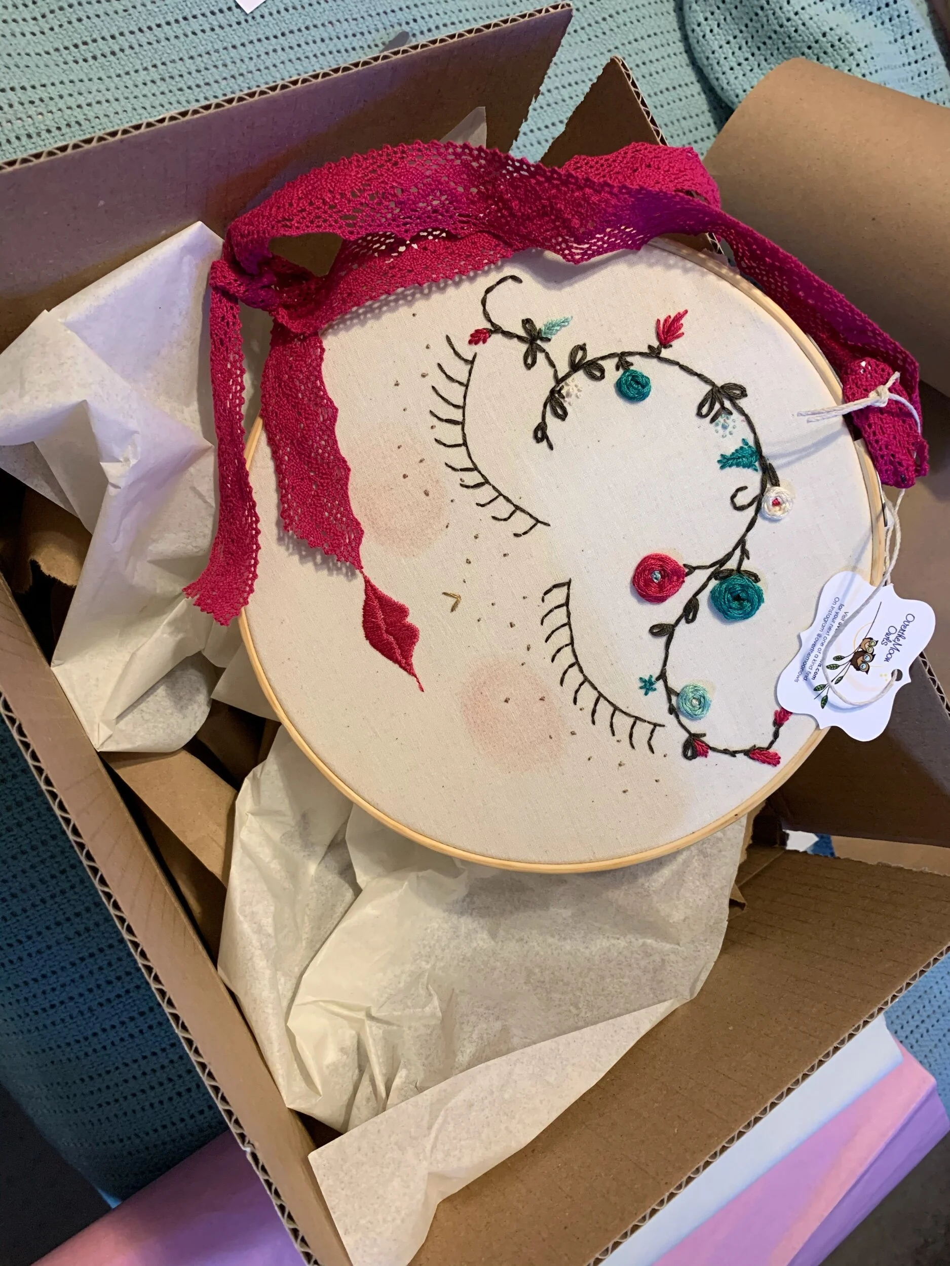Embroidered face design in a hoop with pink lace ribbon, packaged in a cardboard box with tissue paper.