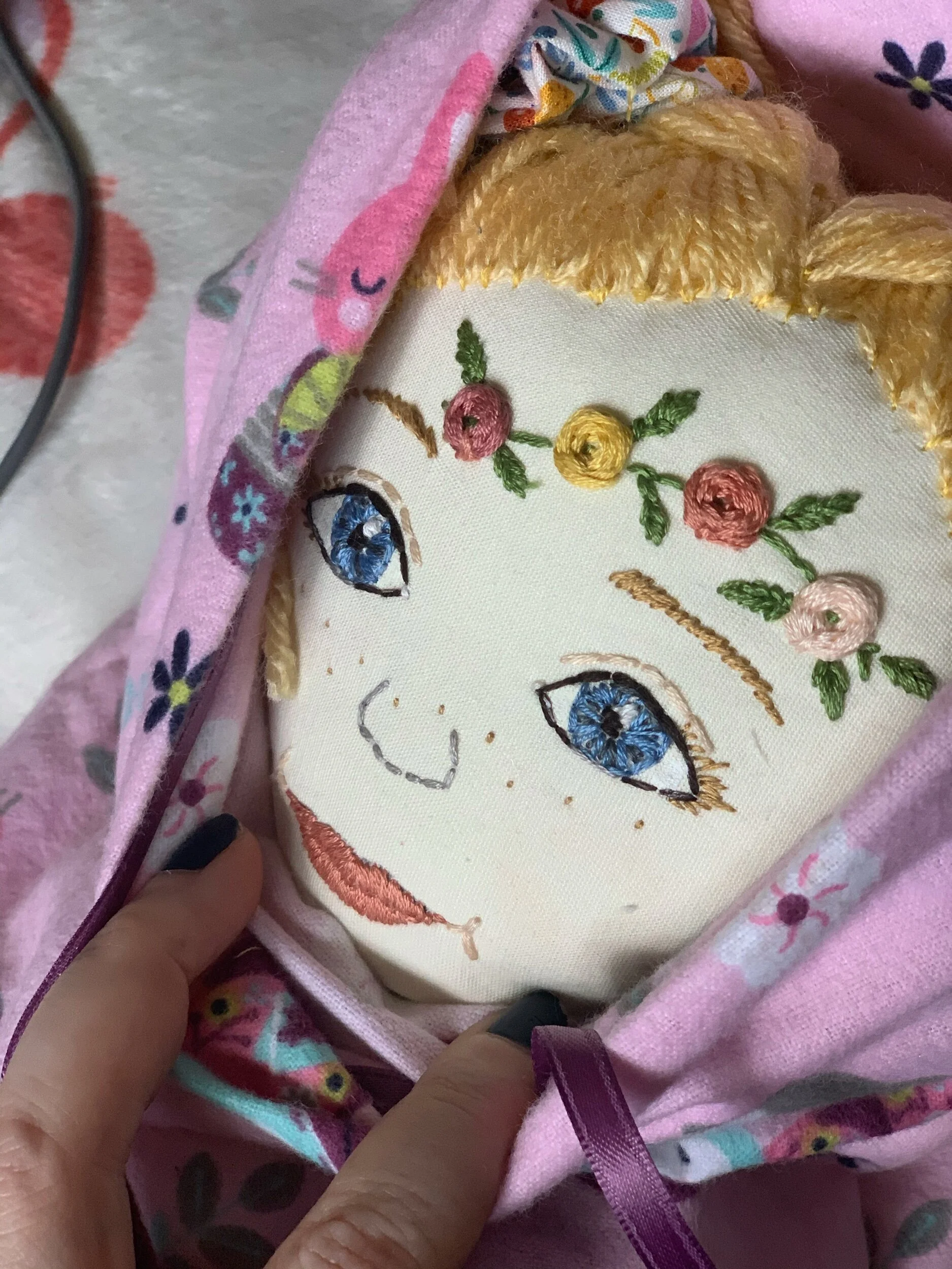 Close-up of an embroidered face on a fabric doll with blue eyes, flower patterns on the forehead, and wrapped in pink fabric.