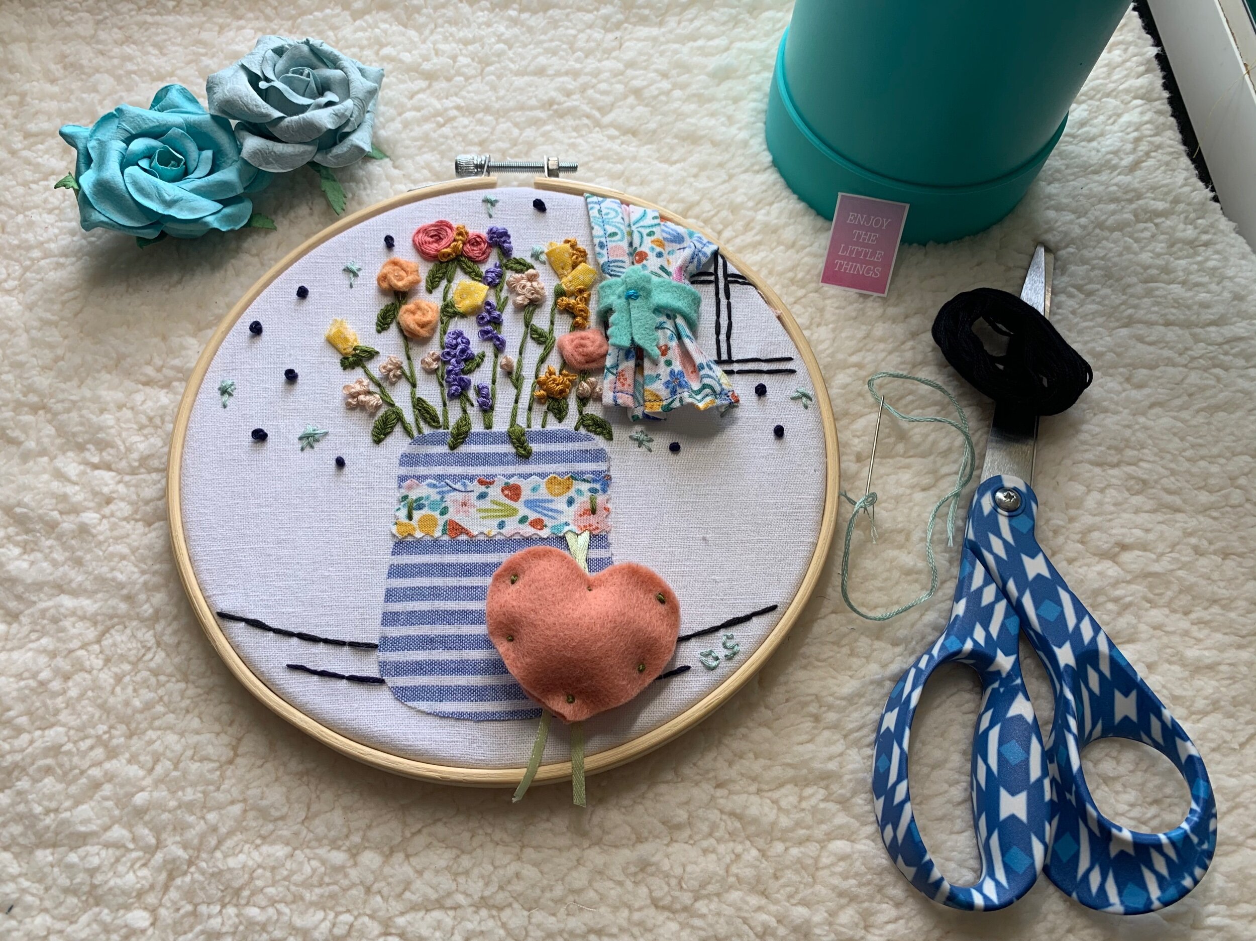 Embroidery hoop with a floral design, scissors, thread, and fabric flowers on a textured surface.