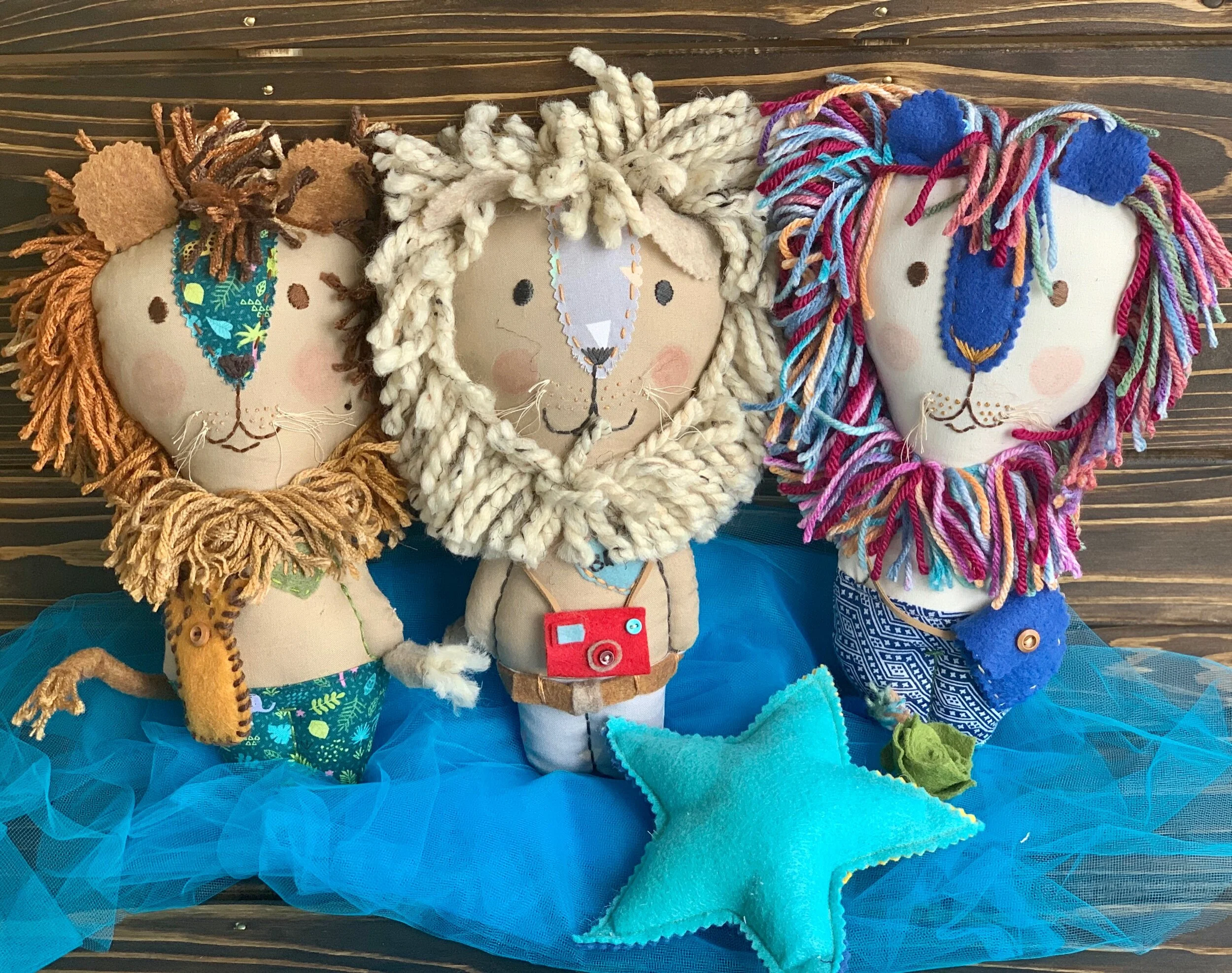 Three plush toy lions with colorful yarn manes and detailed costumes, sitting on a blue tulle fabric with a stuffed blue star in front.