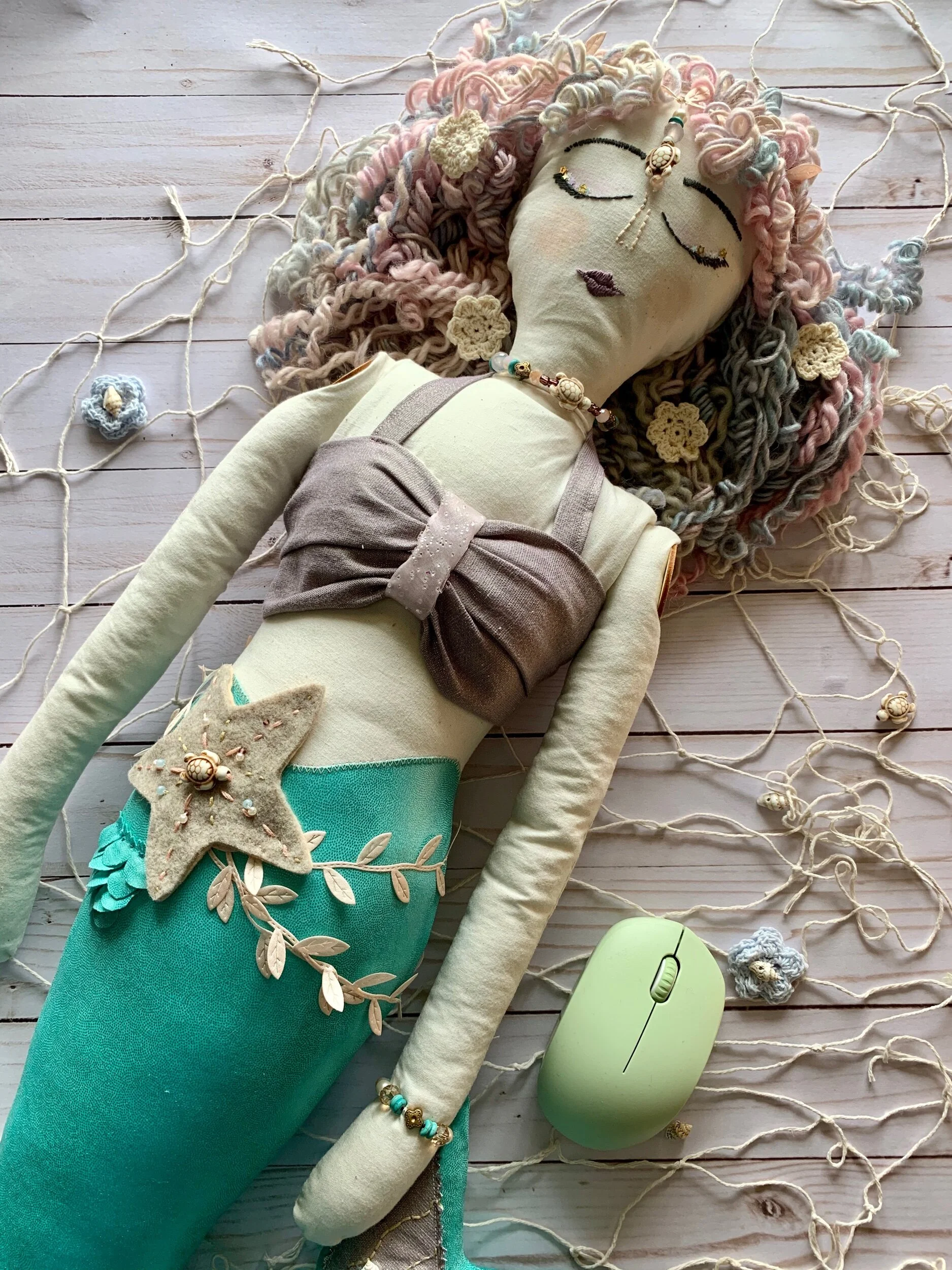 Homemade cloth mermaid doll with colorful yarn hair, decorative beads, and a felt star; green wireless computer mouse nearby on a wooden surface.