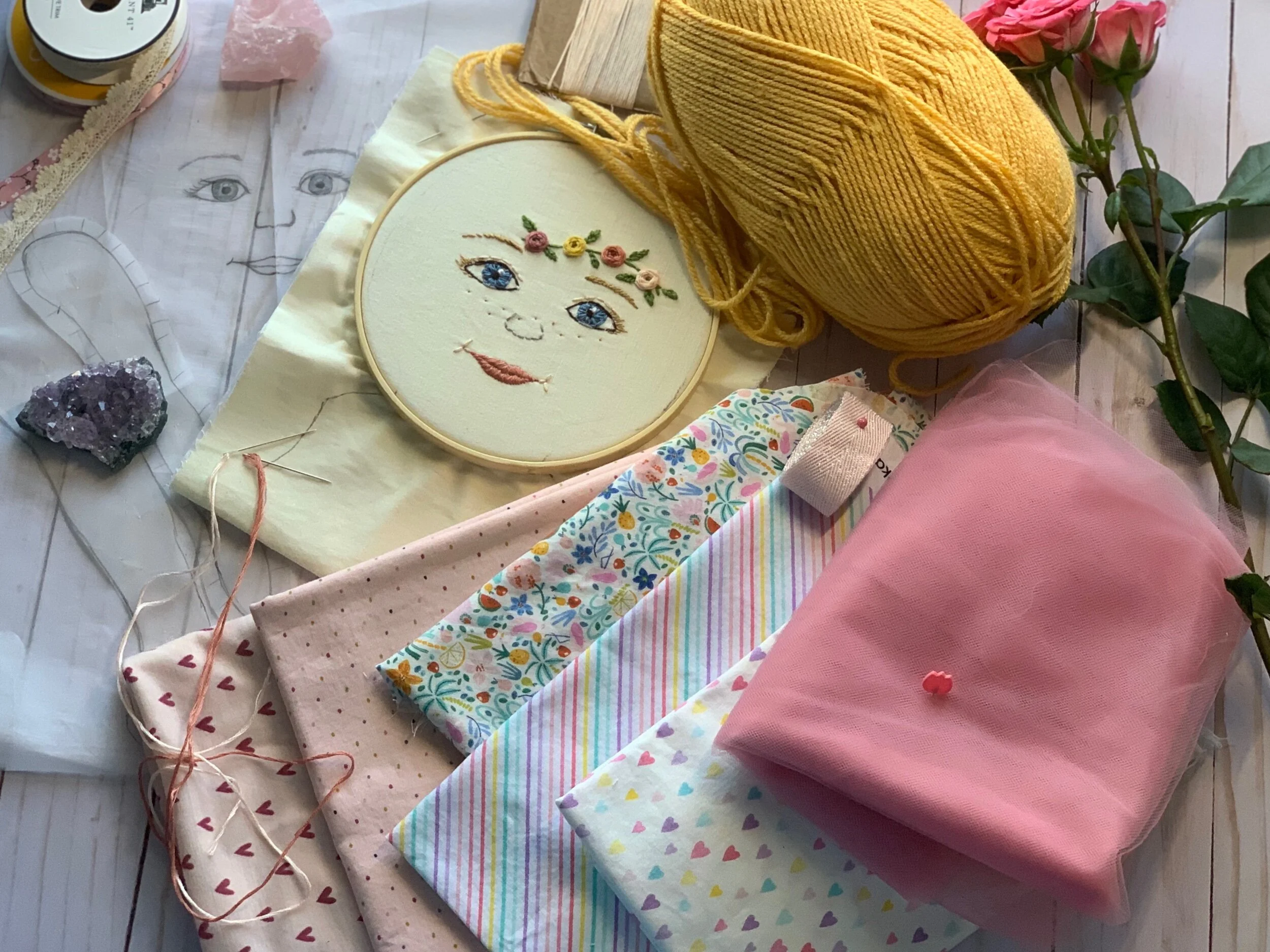 Embroidery and craft supplies, including a hoop with a face design, yellow yarn, fabric swatches with various patterns, a rose, and a piece of amethyst.