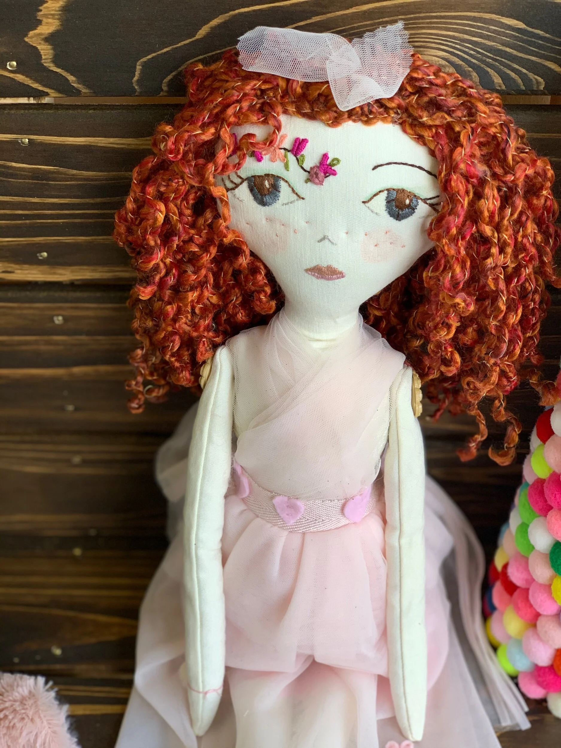 Handmade cloth doll with curly red hair, pink dress, and floral embroidery, sitting on a wooden surface.