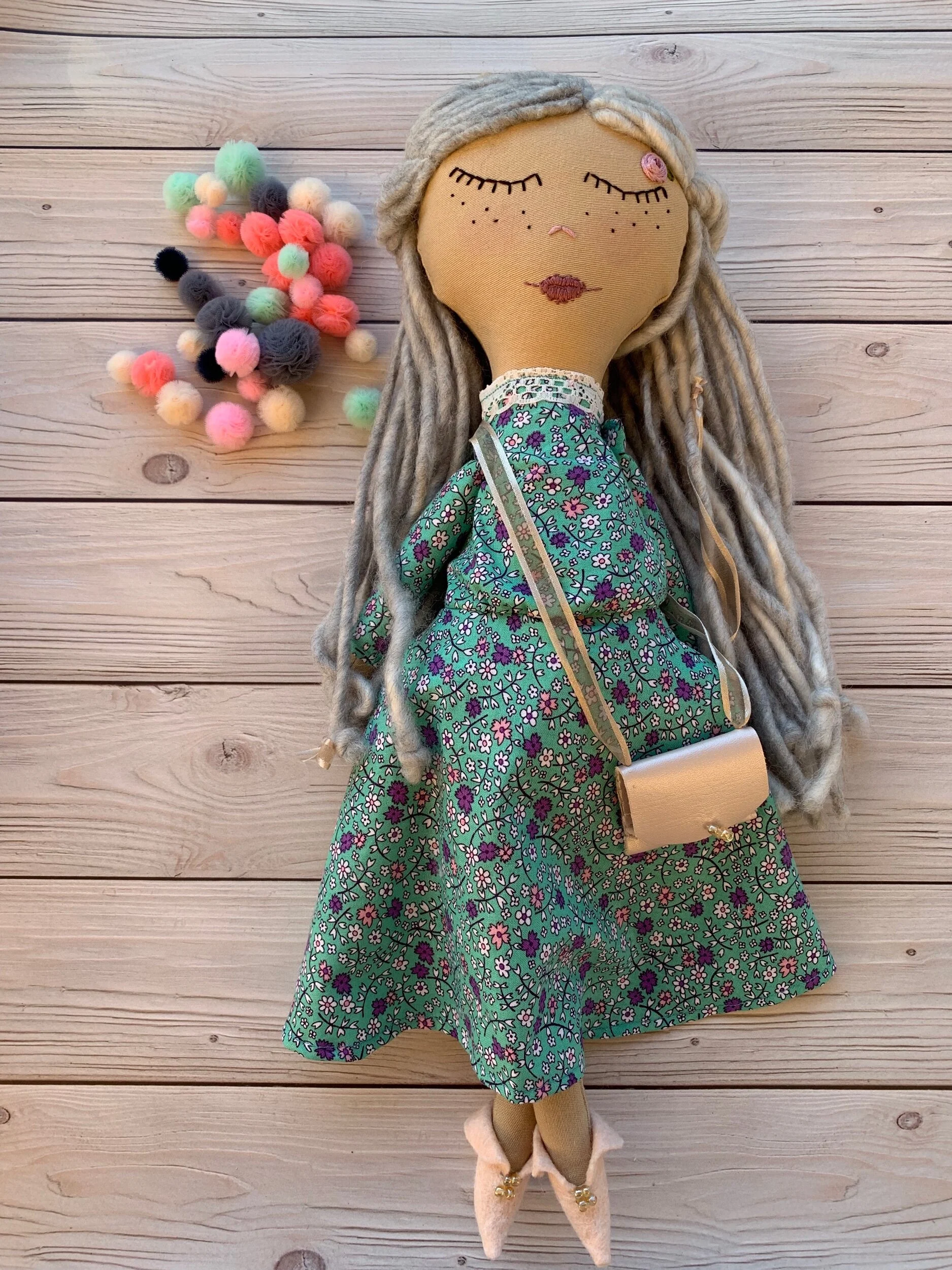 Handmade cloth doll with long gray hair, wearing a green floral dress and holding a small beige purse, lying on a wooden surface next to colorful pom-pom clusters.