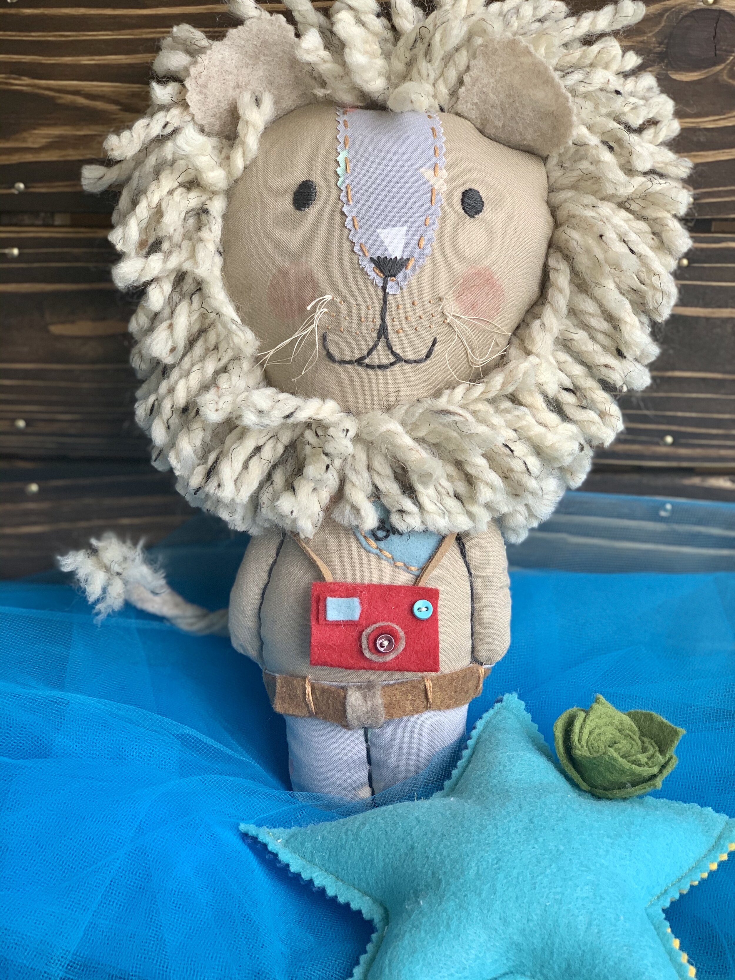 Stuffed toy lion with yarn mane, felt camera, standing on blue fabric.