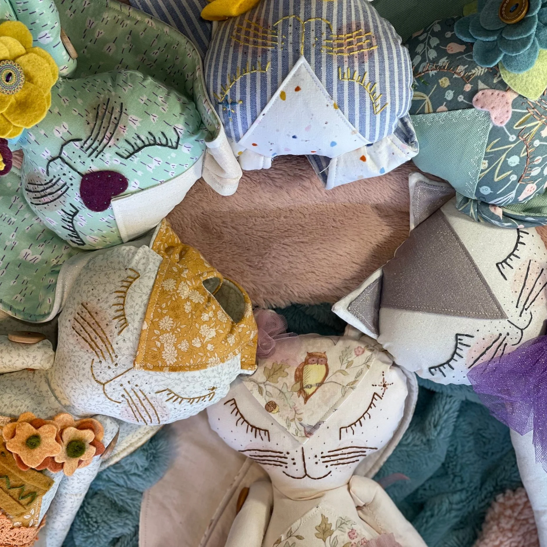 Assorted handmade fabric cat plushies with colorful patterns and floral details arranged in a circle on a soft surface.