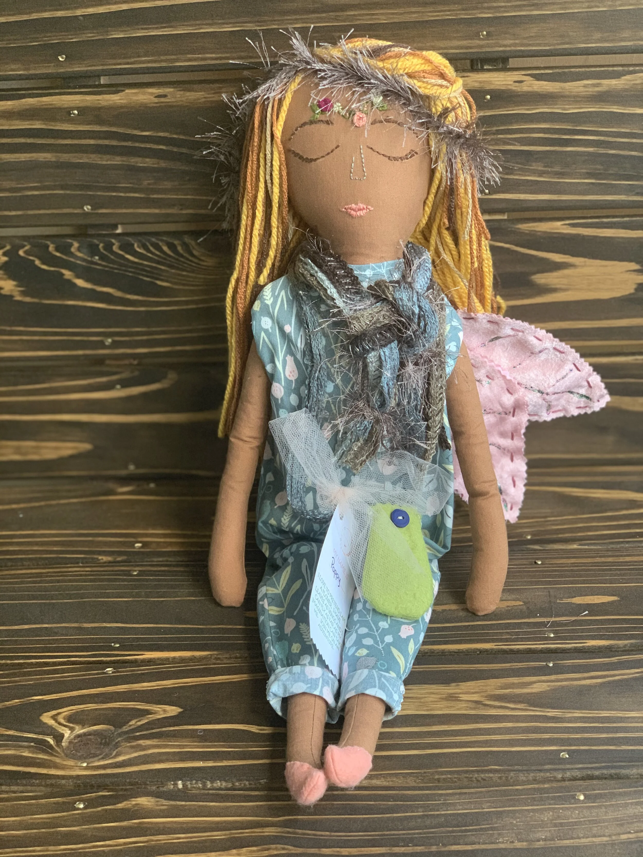 Handmade cloth doll with brown skin tone, multicolored yarn hair, wearing a blue floral jumpsuit, pink shoes, a fabric scarf, and pink wings. The doll is sitting against a wooden background.