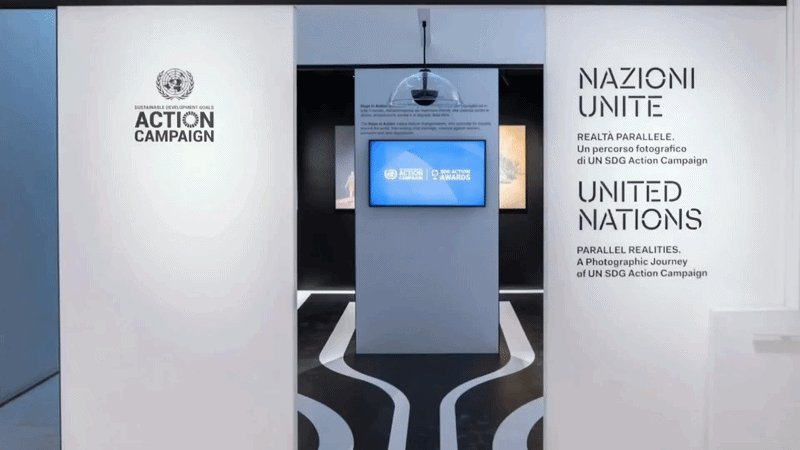 A metallic sculpture of a revolver with a hand gripping it, mounted on a black stand that reads 'NON-VIOLENCE - In memory of John Lennon.' In the background, there is a large pink flower-shaped artwork on the wall.