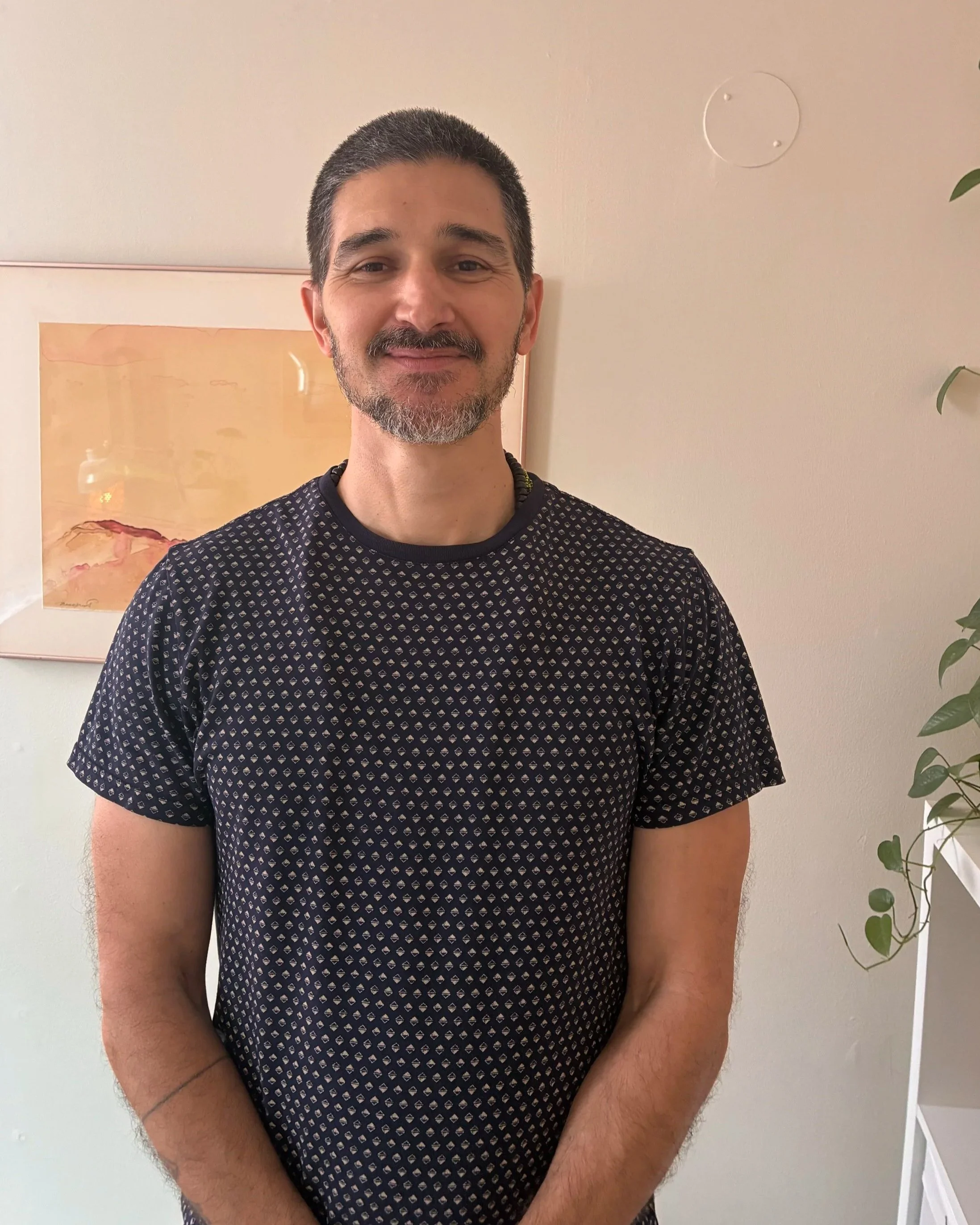 A man with short gray hair and a beard, wearing a black shirt with small geometric patterns, smiling in front of a light-colored wall with framed artwork and a leafy plant.