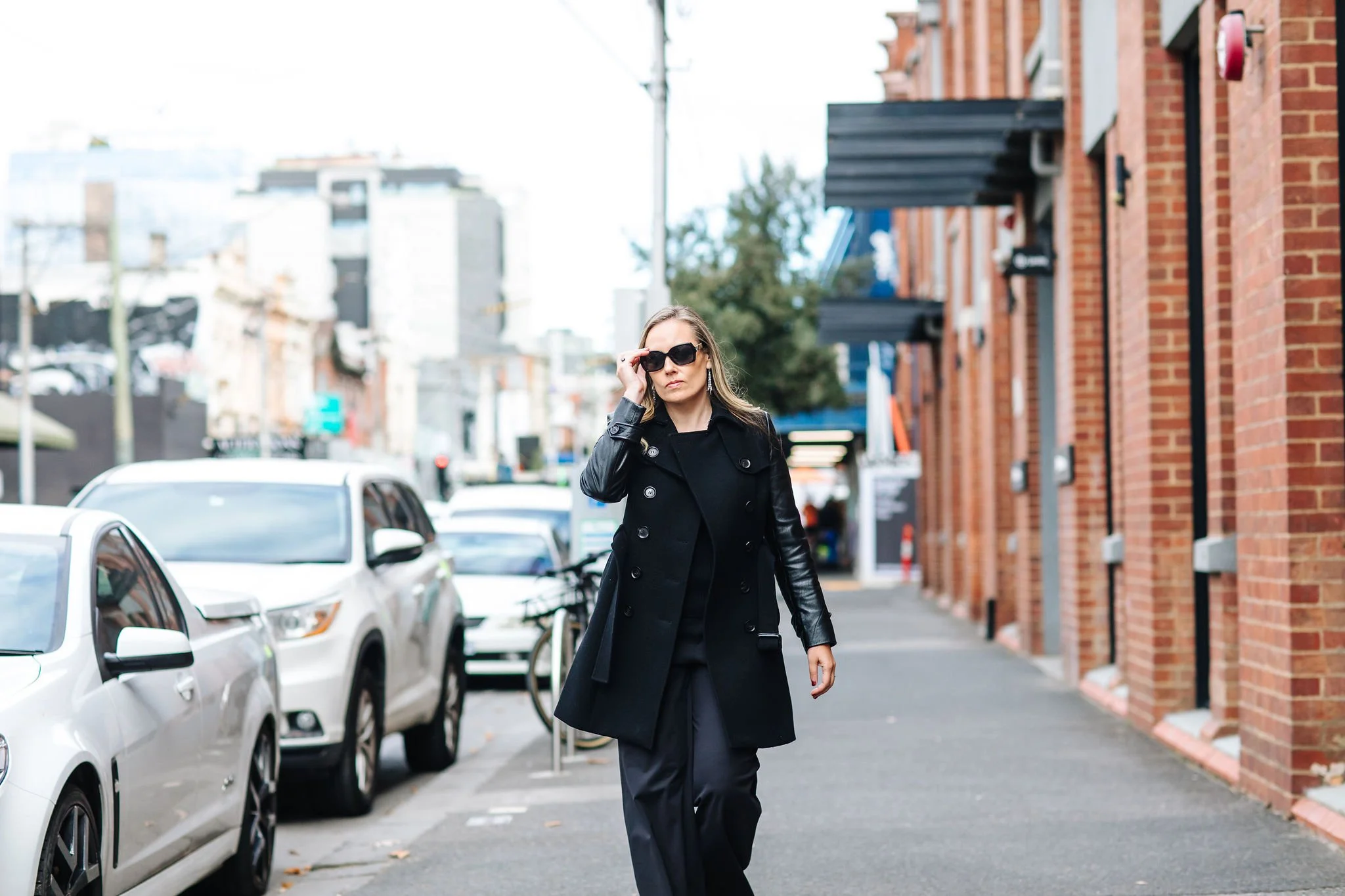 rebecca kartel walking on sidewalk wearing all black and shades with brick wall background and cars parked