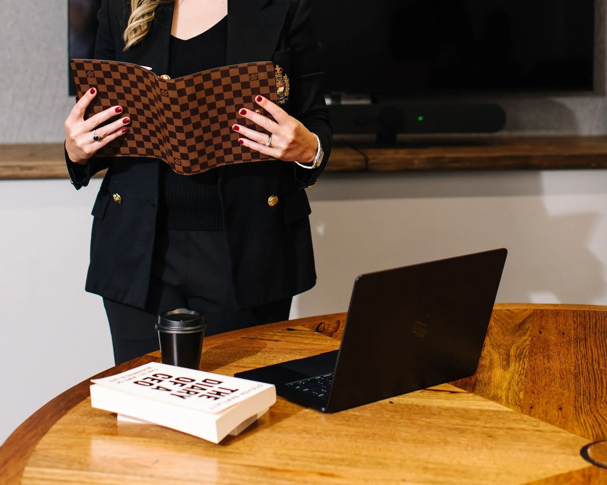 A woman with blonde hair, wearing a black blazer, sitting at a wooden conference table with a laptop and sunglasses, in a modern office setting.