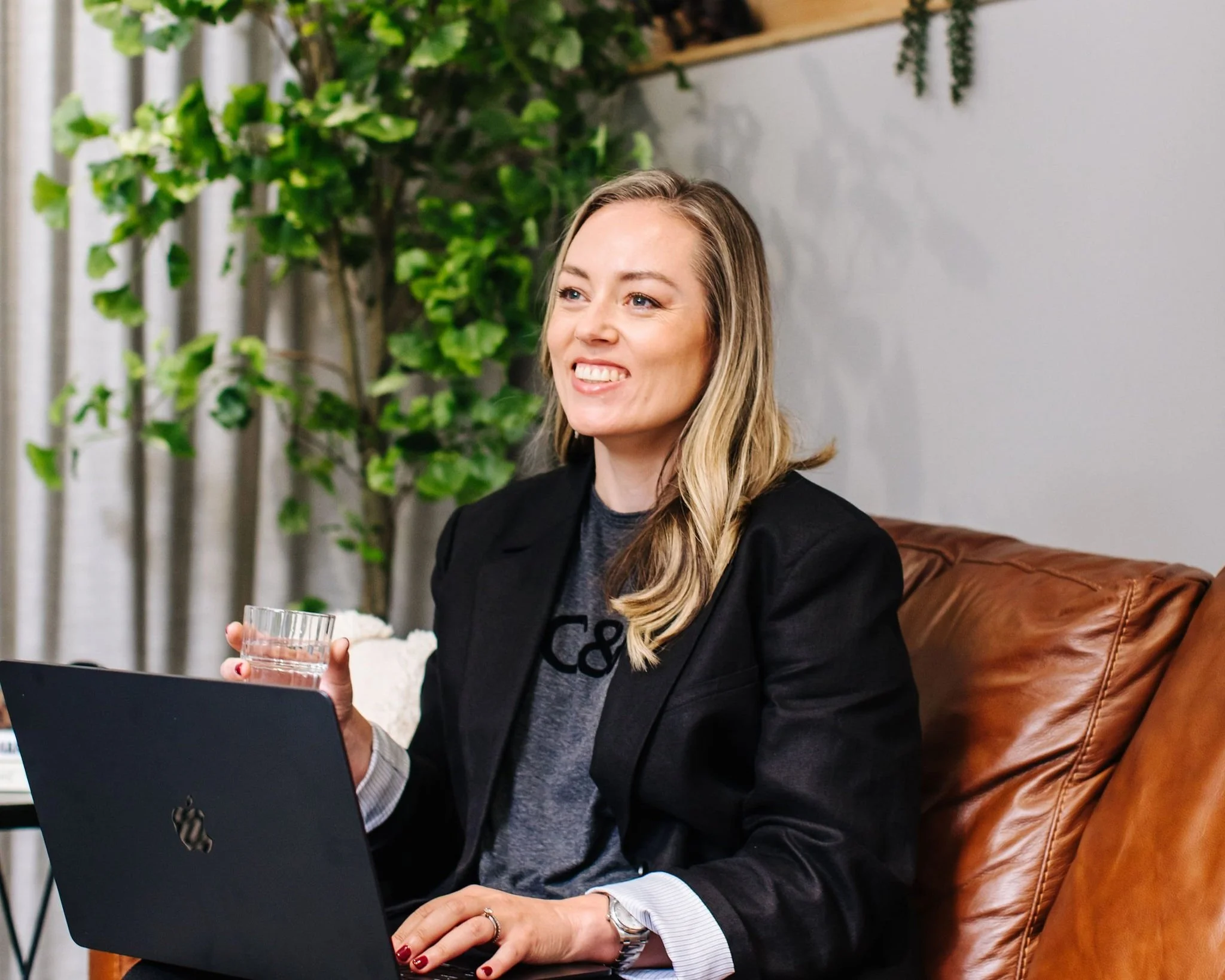 A woman with blonde hair in a black blazer holding a glass of whiskey while working on a laptop, seated on a multicolored modern chair in front of a wooden slat wall.