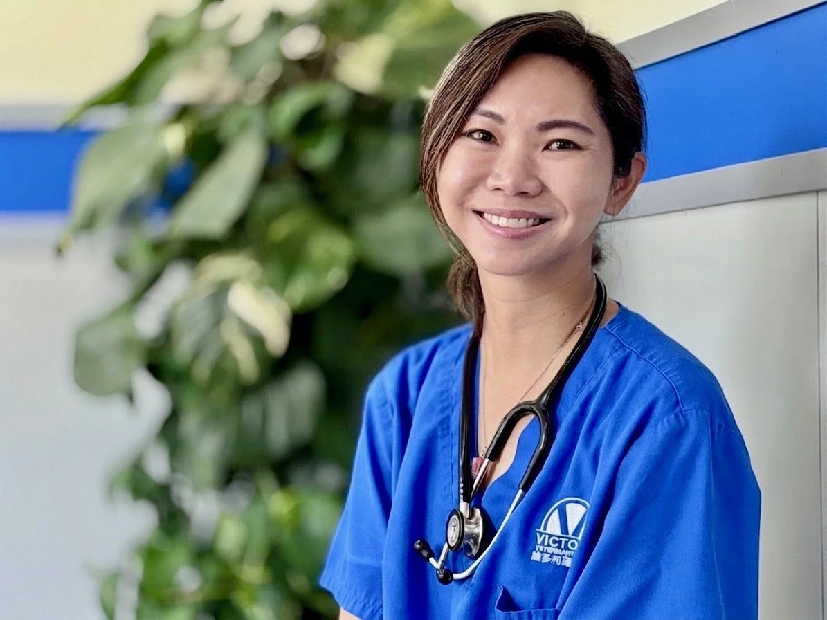 Dr Joyce wearing blue scrubs and a stethoscope around her neck, sitting next to a wall with green plants in the background.