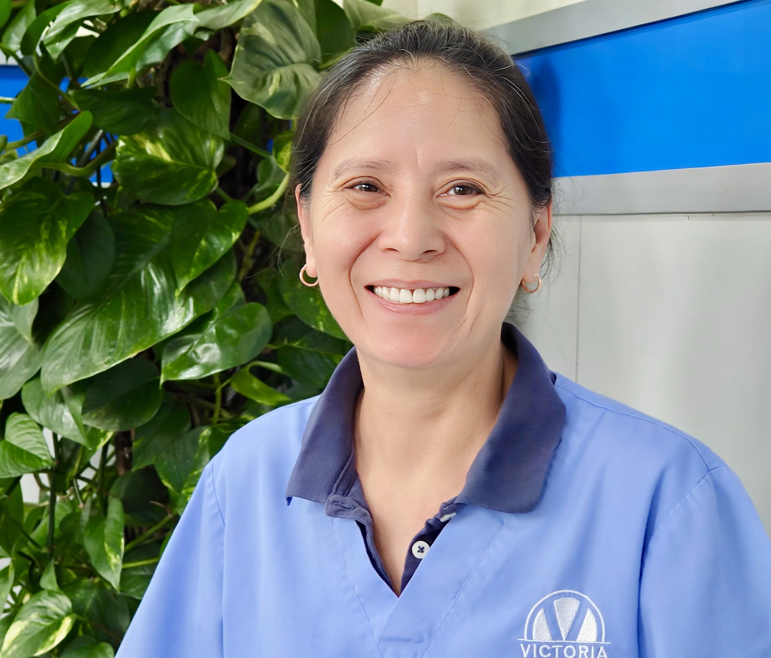 Close-up of a smiling woman, Mandy,  in a blue collared shirt with a Victoria logo, standing outdoors next to green leafy plants.