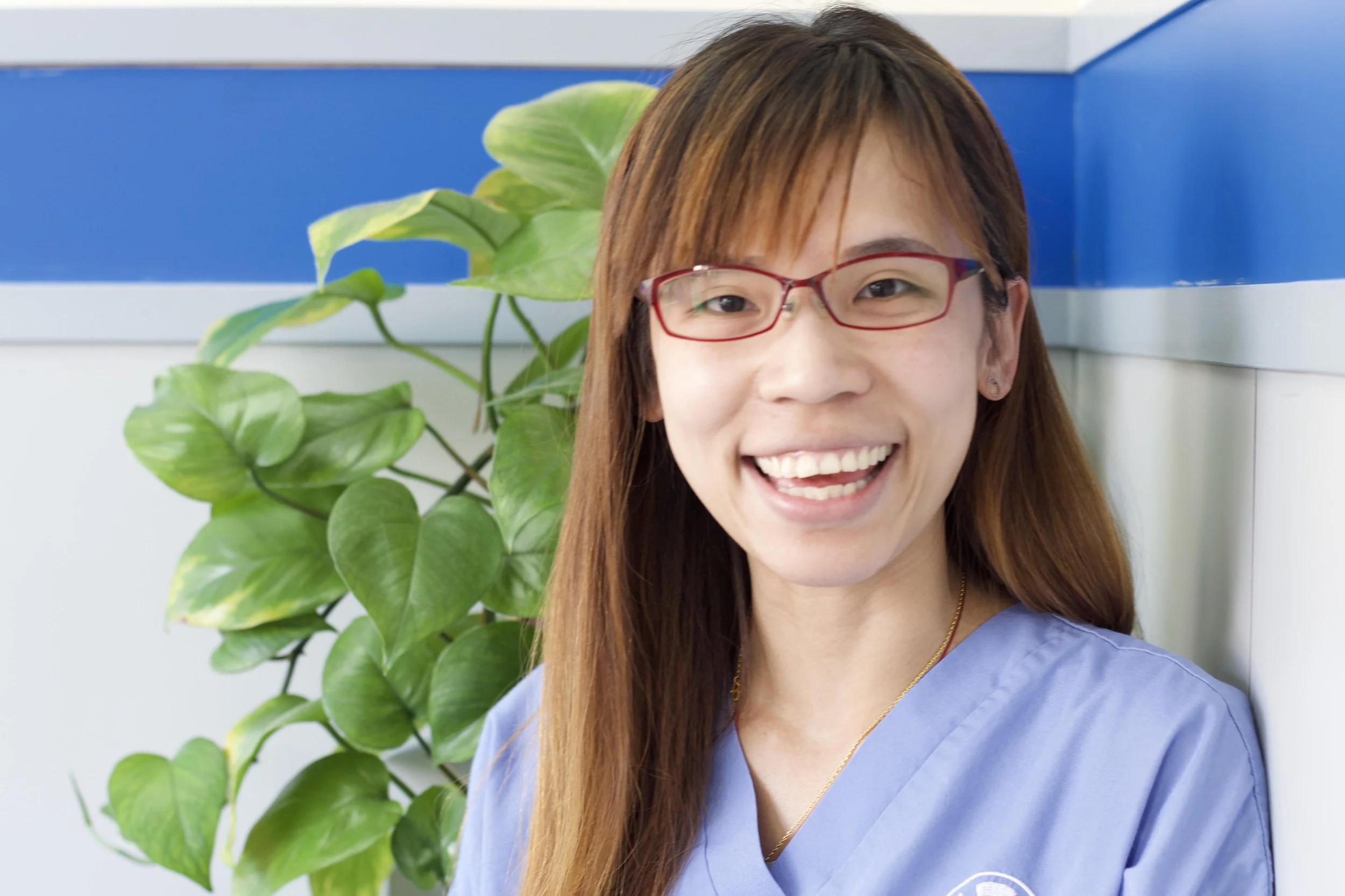 Smiling young woman, Cholas, with red glasses and brown hair wearing light blue scrubs, standing in front of a leafy green plant and a blue wall.
