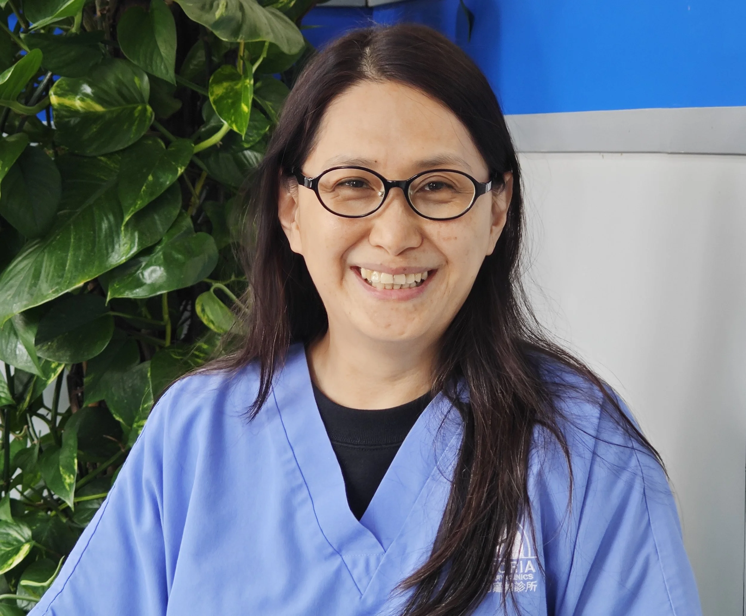 Close-up of a smiling woman, Carol, with glasses, long dark hair, wearing a blue medical scrub top, standing outdoors with green leafy plants in the background and a blue and white wall.