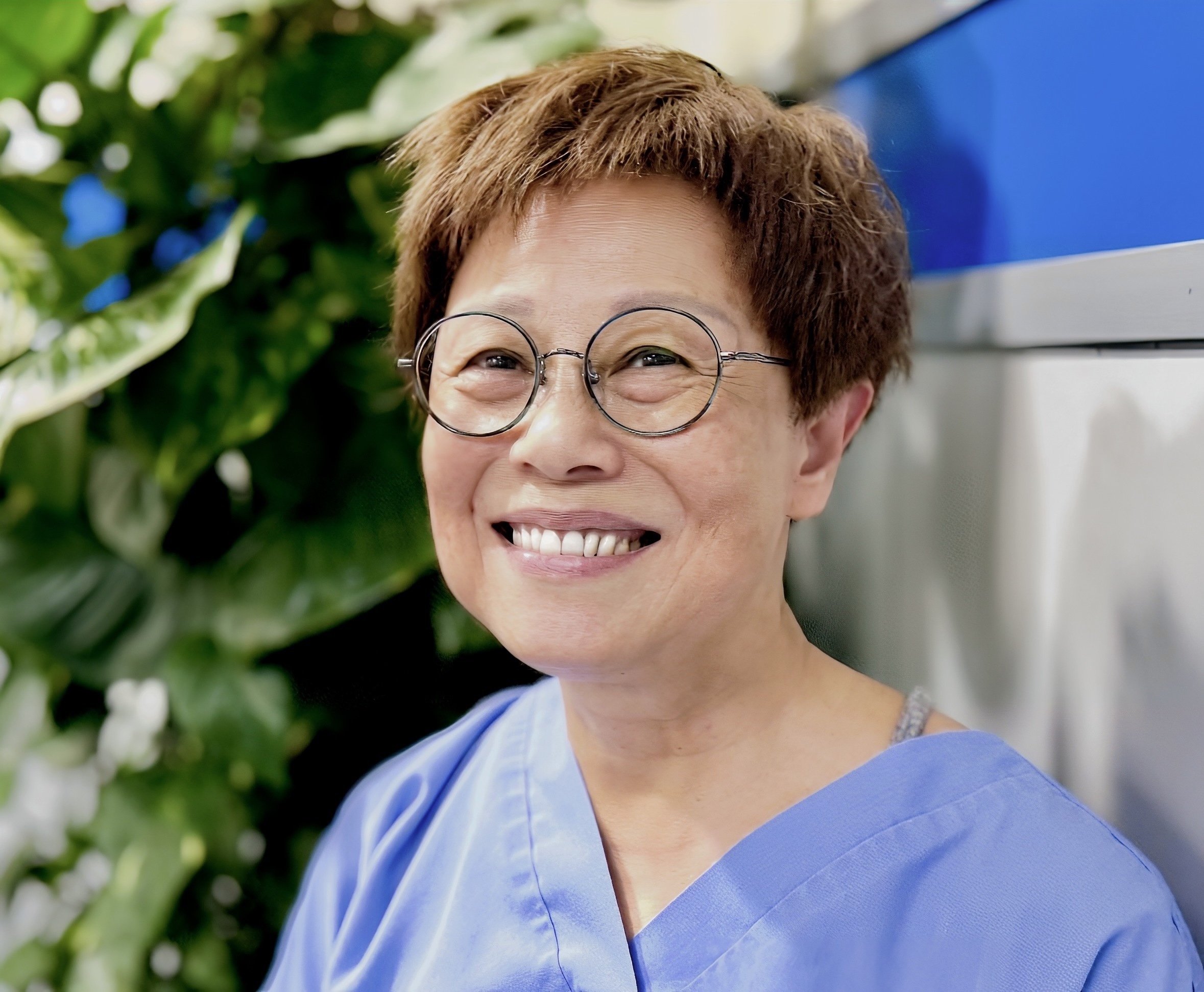 Smiling woman, Fanny,  with short brown hair, wearing round glasses and a blue top, standing next to green leafy plants.