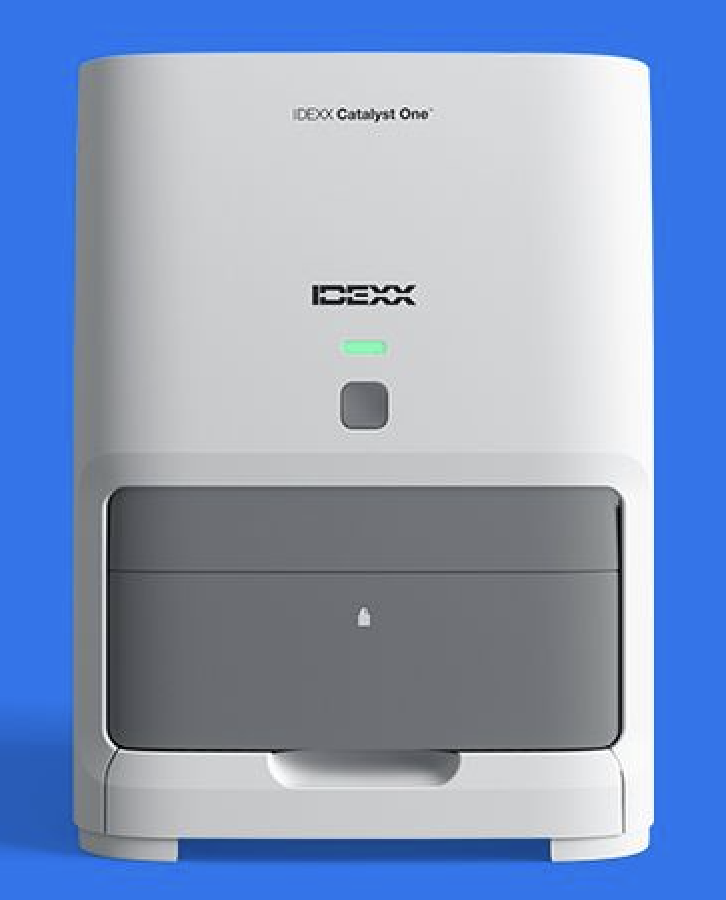 A white IDEXX Catalyst One laboratory analyzer with a grey drawer, placed against a bright blue background.