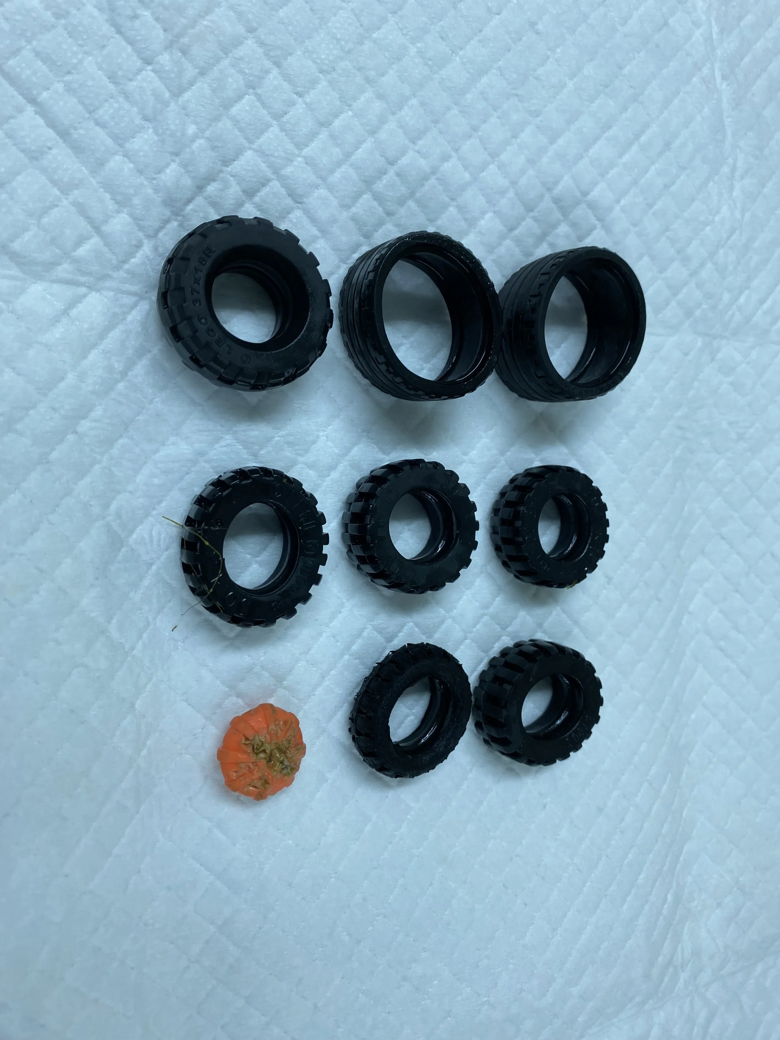 eight black rubber tires arranged on a white quilted surface, with a small orange object at the bottom left corner.