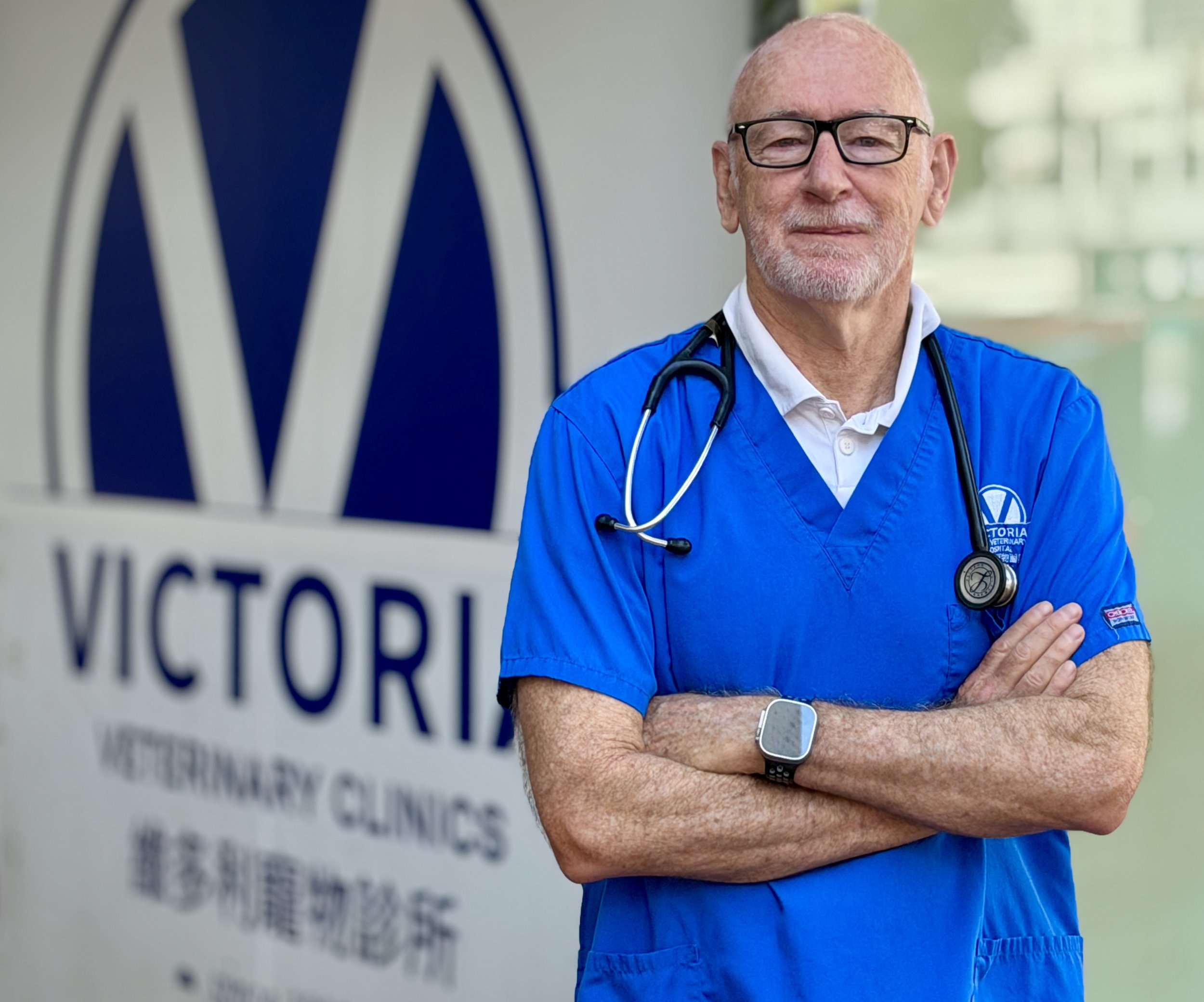 Dr Ken Thorley wearing glasses and blue scrubs with a stethoscope around his neck, standing with arms crossed in front of Victoria veterinary clinic.
