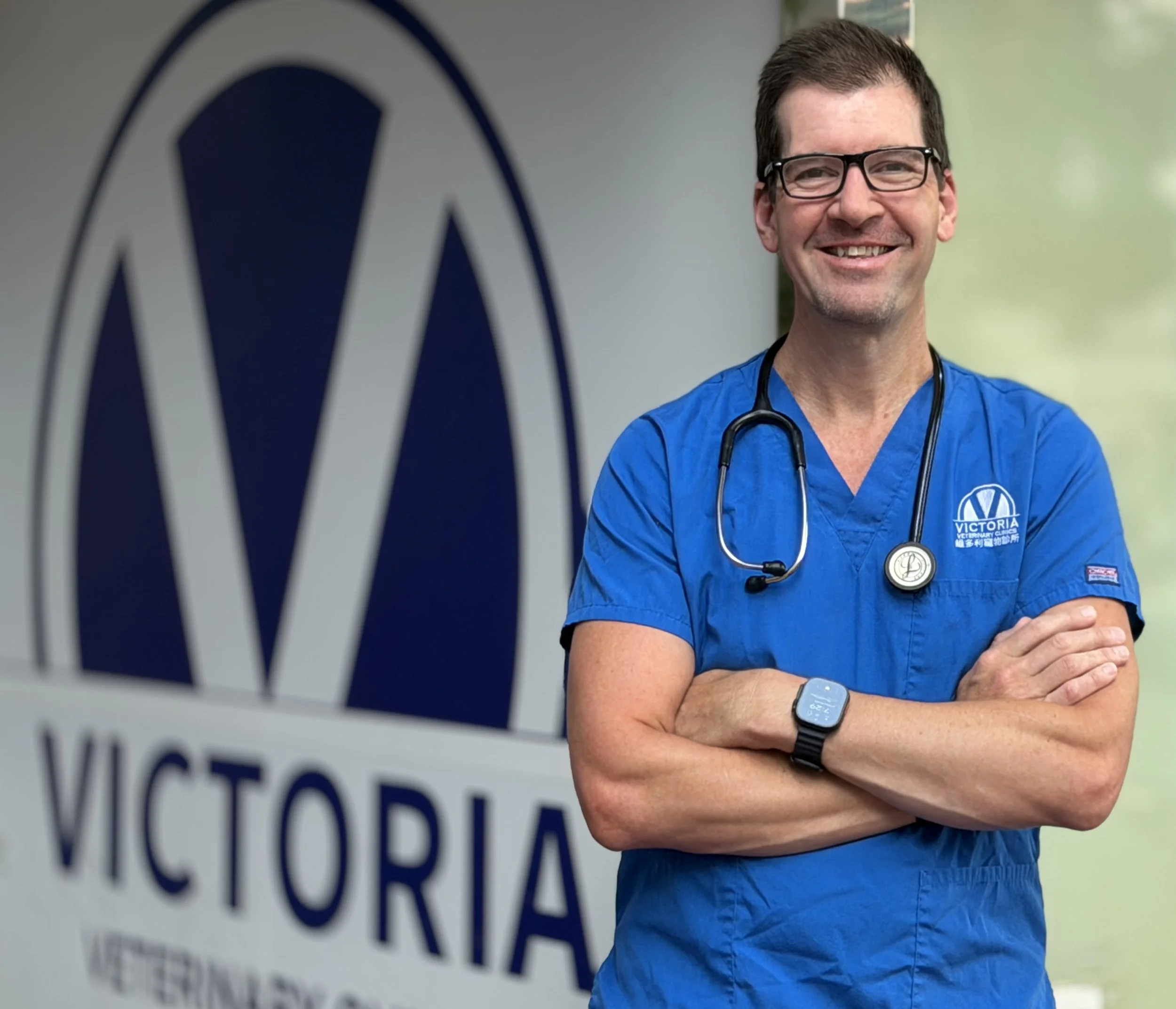 Dr Chris wearing glasses and blue scrubs with a stethoscope around his neck, standing with arms crossed in front of a sign that reads 'Victoria Veterinary Clinic.'