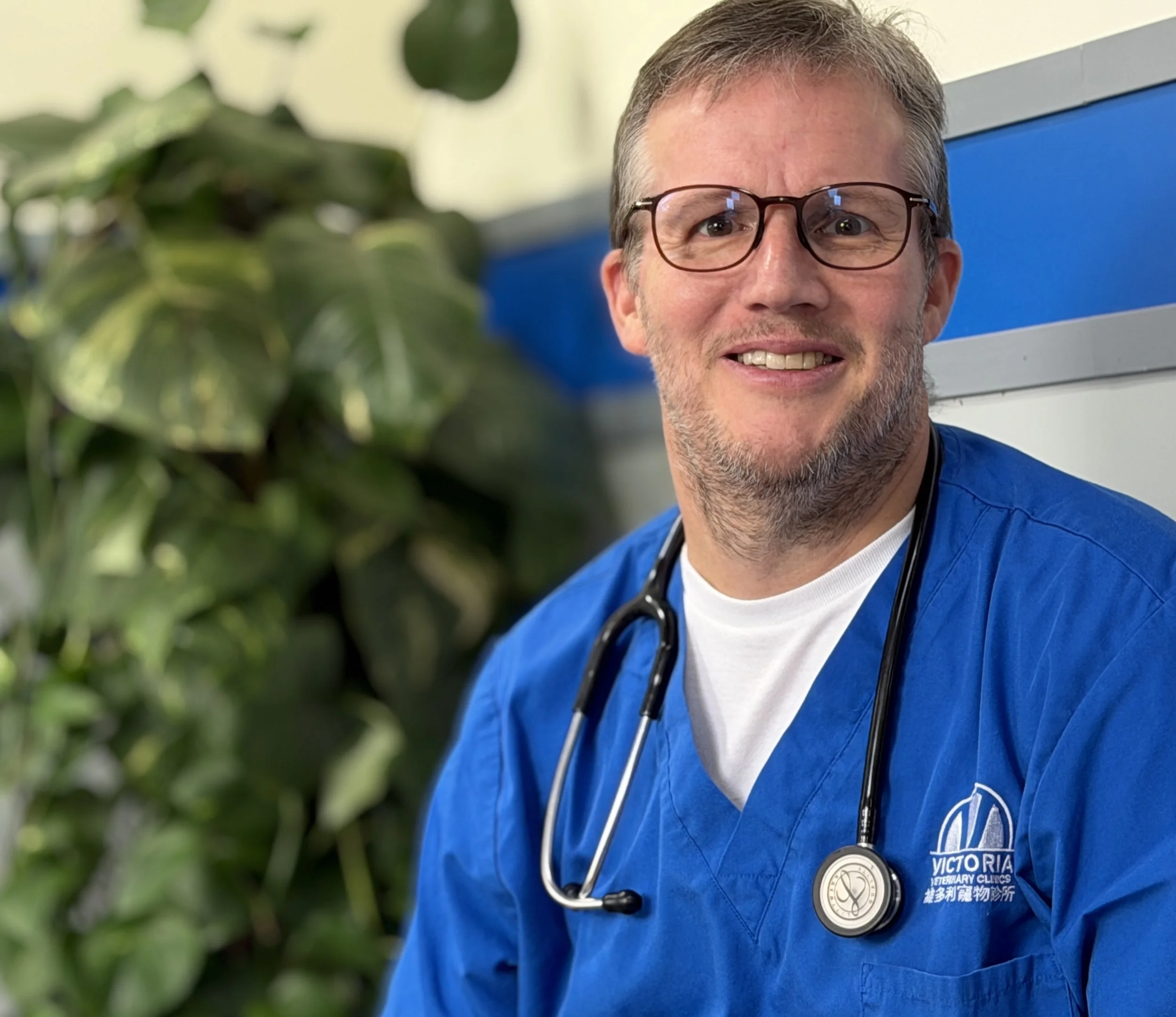 Dr Jeremy wearing glasses and blue scrubs with a stethoscope around his neck, standing in front of a blue and gray background, with some green foliage in the foreground.