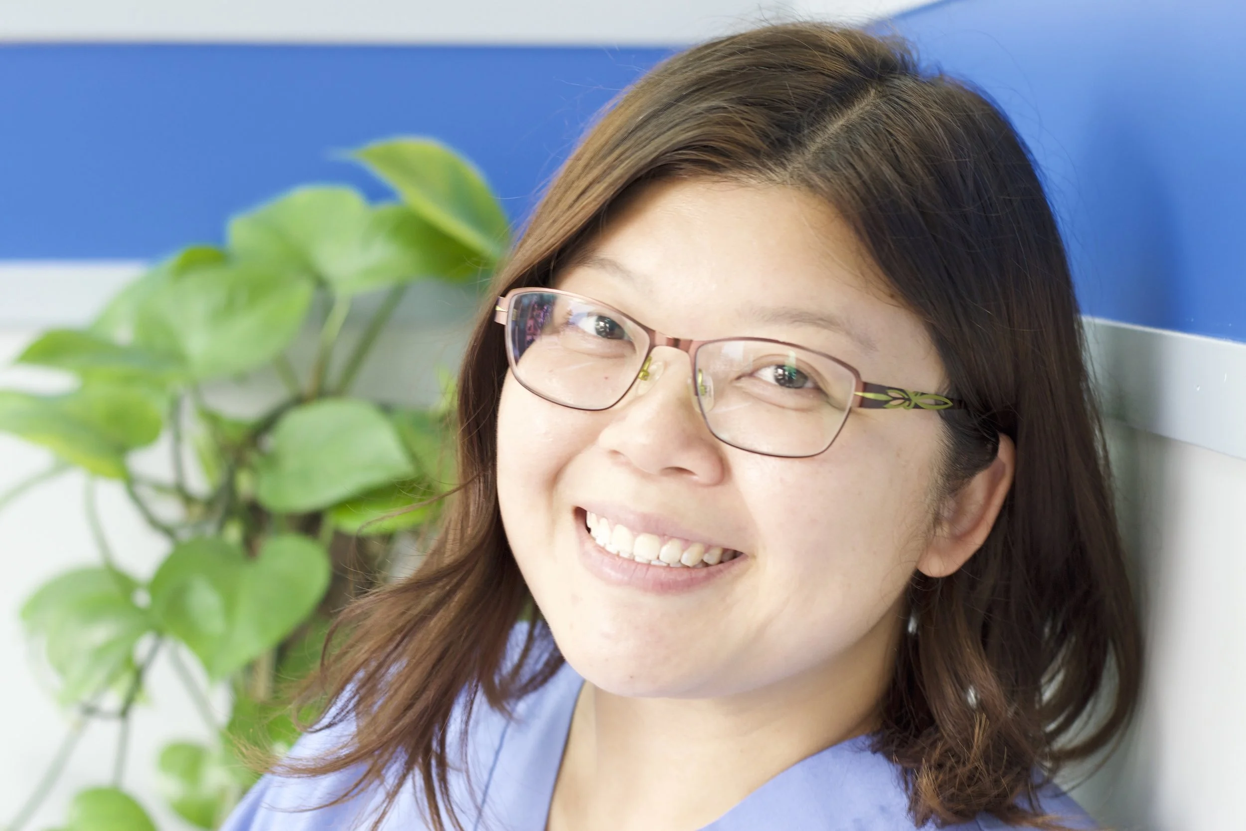 Close-up of a woman, Wing, with brown hair and glasses smiling, with green leaves and a blue wall in the background.
