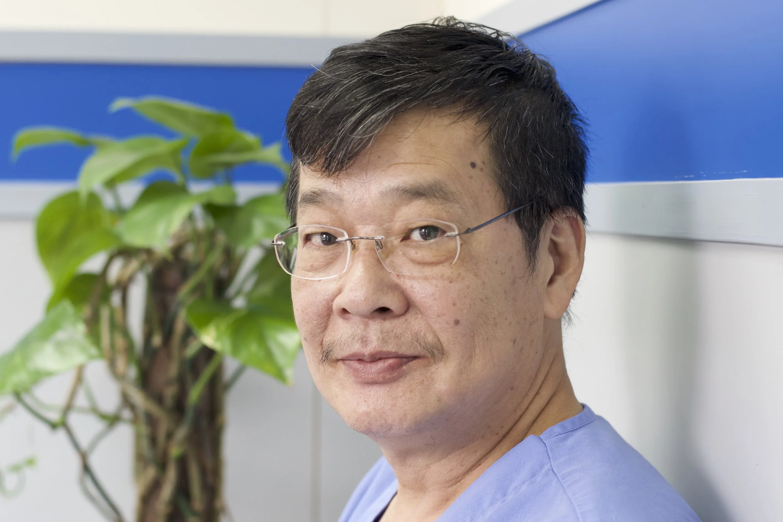 Close-up of a man, Patrick, with black hair, glasses, and a light blue shirt, standing next to a green potted plant in an indoor setting.