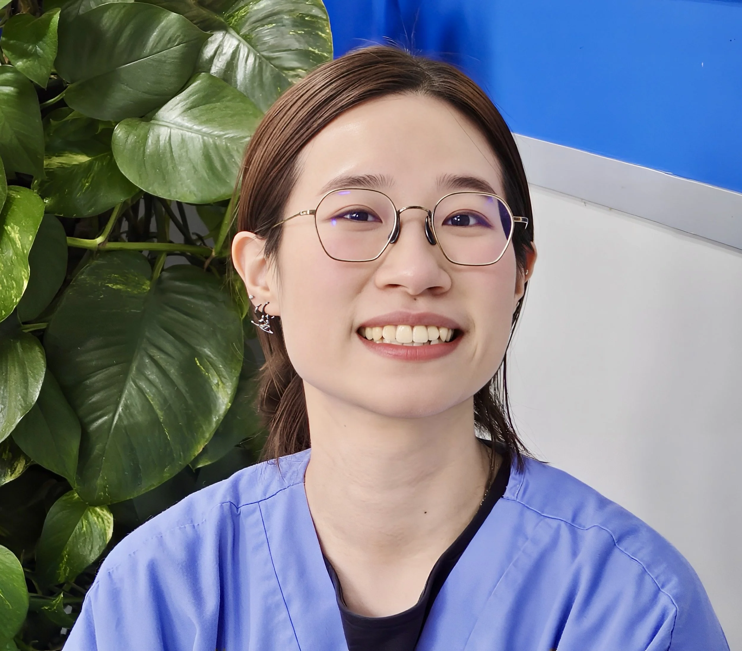 Close-up of a young woman, Gloria,  smiling, wearing glasses with a large green leafy plant behind her on the left and a blue wall with a white trim on the right.