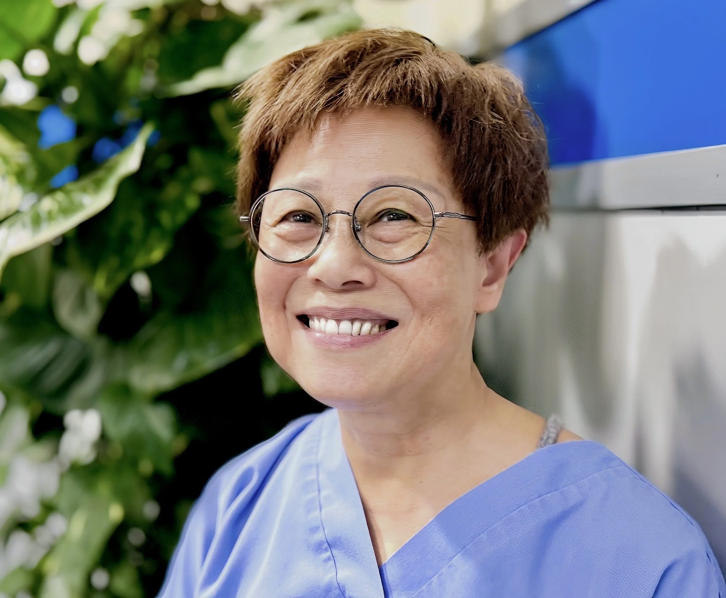 Smiling woman, Fanny,  with short brown hair, wearing round glasses and a blue top, standing next to green leafy plants.