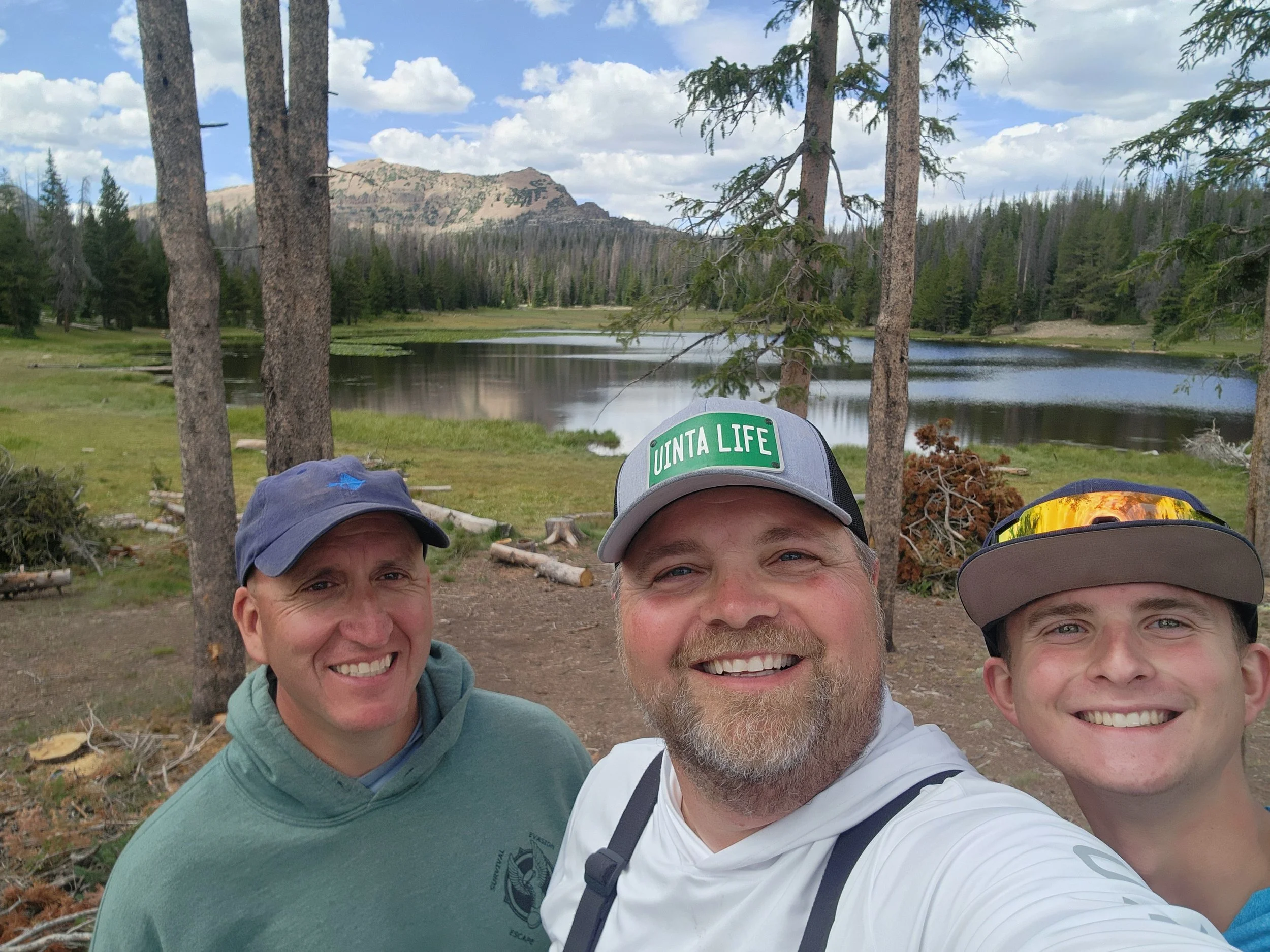 Three men smiling for a selfie outdoors in a forest near a lake, with mountains and a cloudy sky in the background.