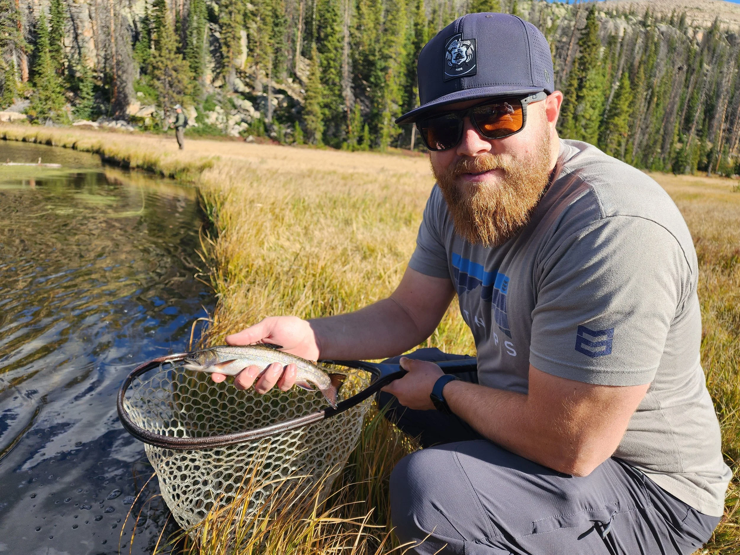 Man with a beard wearing sunglasses, a cap, and a gray t-shirt kneels near a pond and holds a fishing net with a small fish inside.
