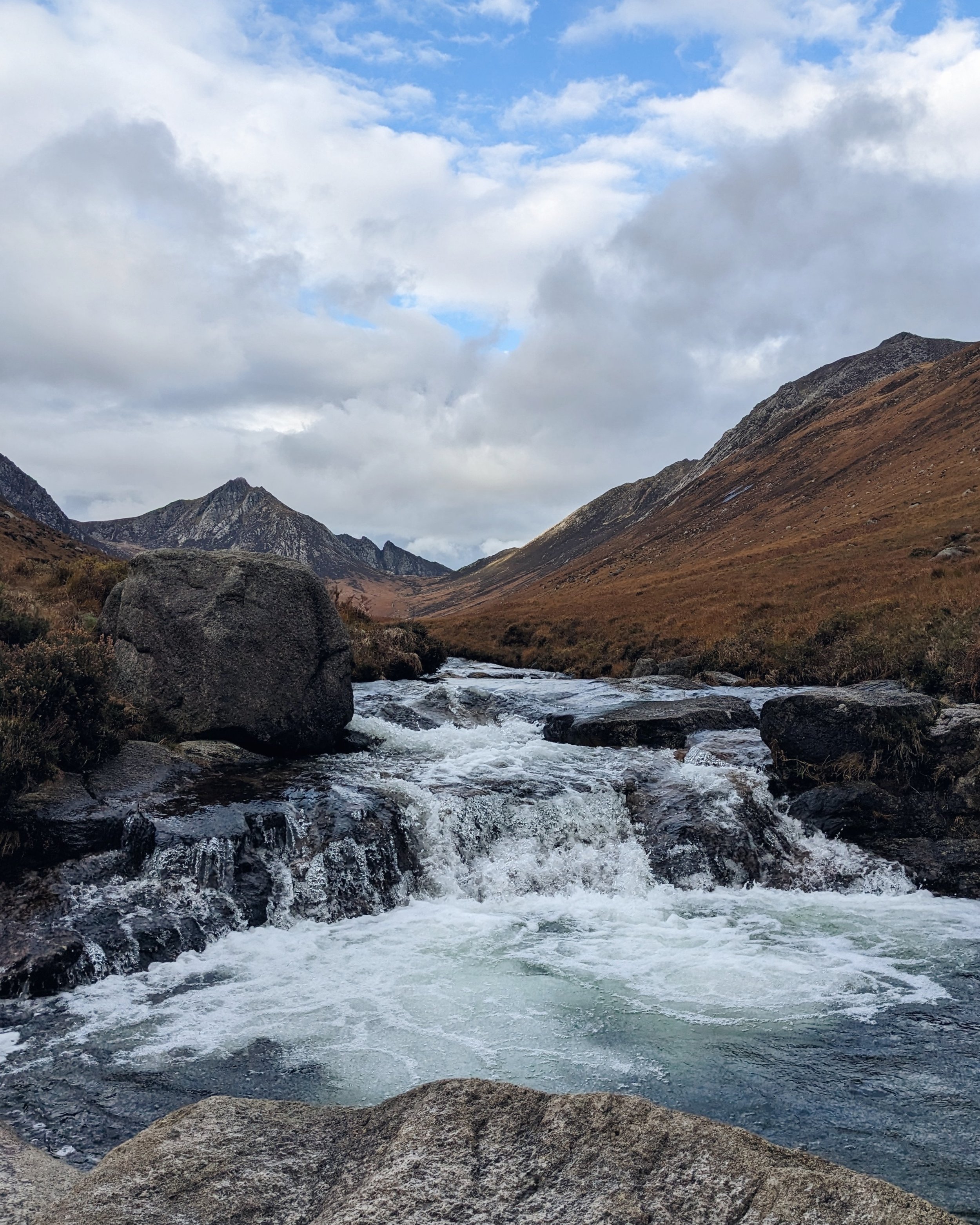 The blue pool in Glen Rosa, a rock pool with mountains behind. The water is grey, reflecting the cloudy sky and the hillsides on the glen are dark orange and brown in late autumn.