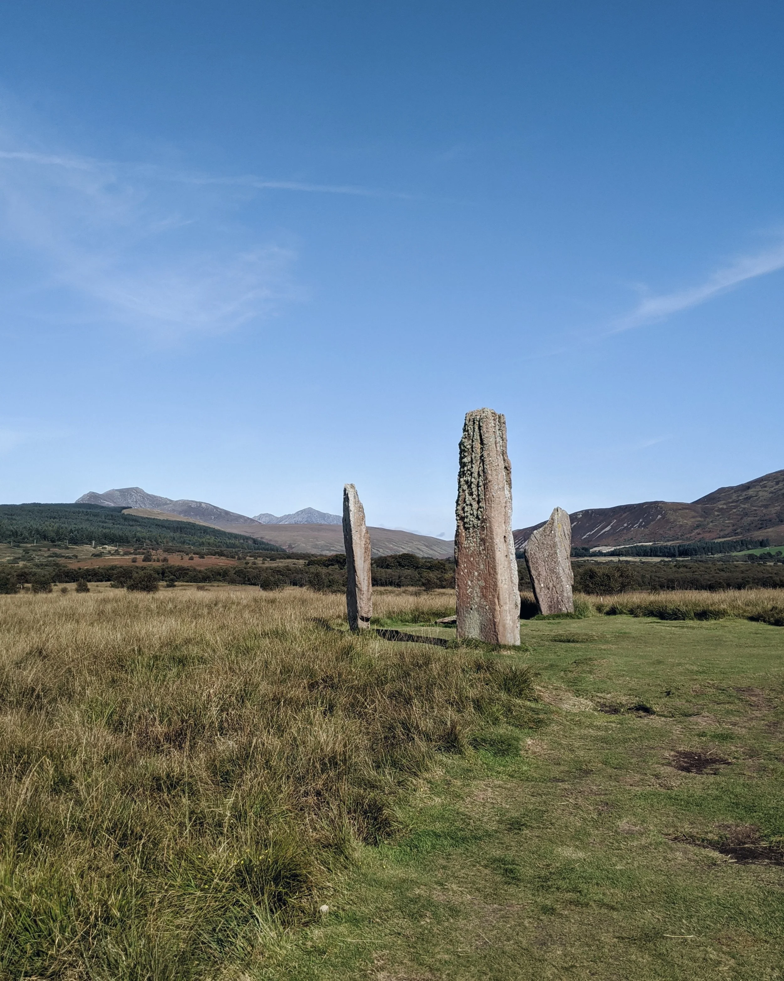 Three standing stones in a grassy field with hills and mountains in the distance.