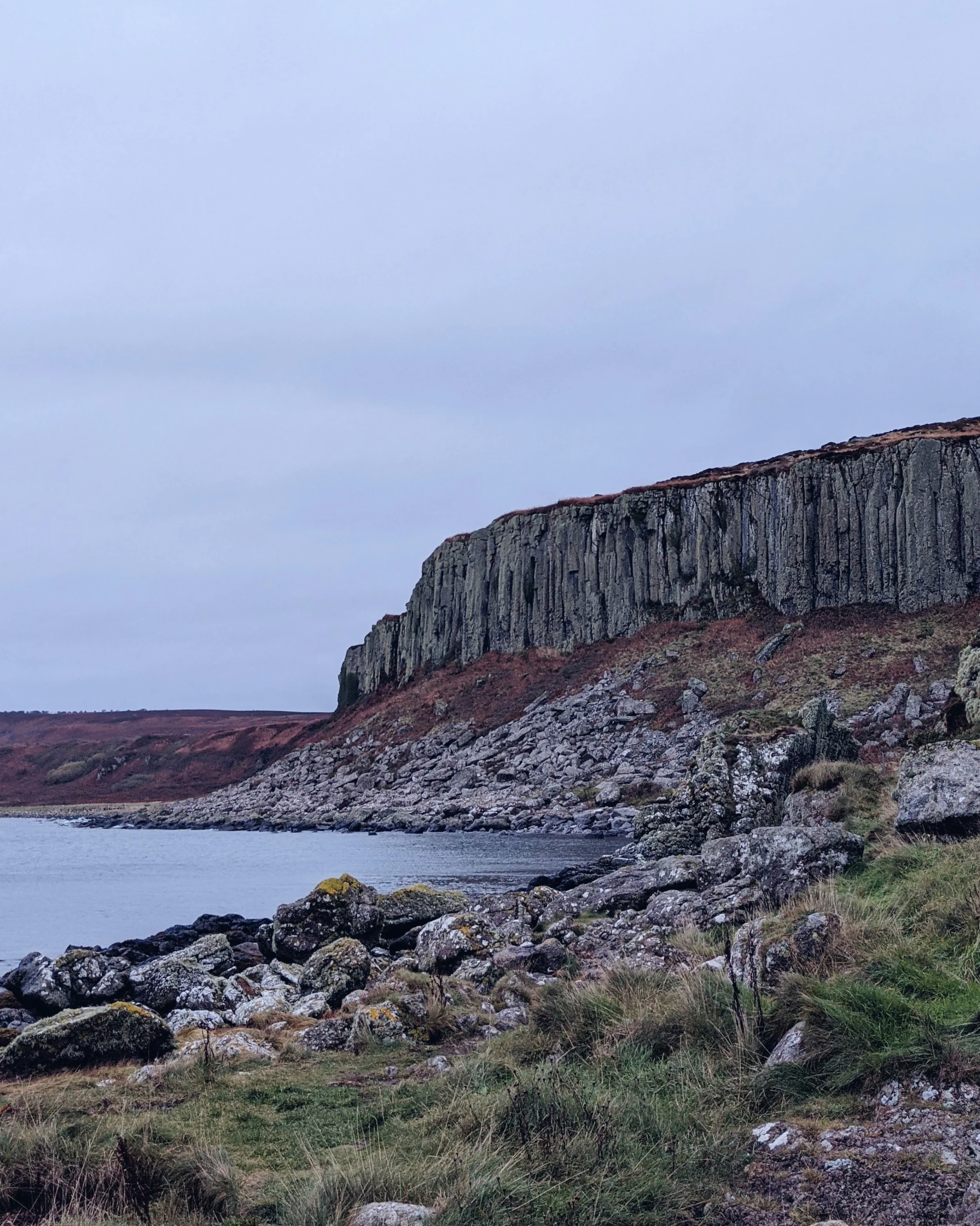A steep cliff face sits above a rocky beach on a cloudy day. The sea and sky are grey.