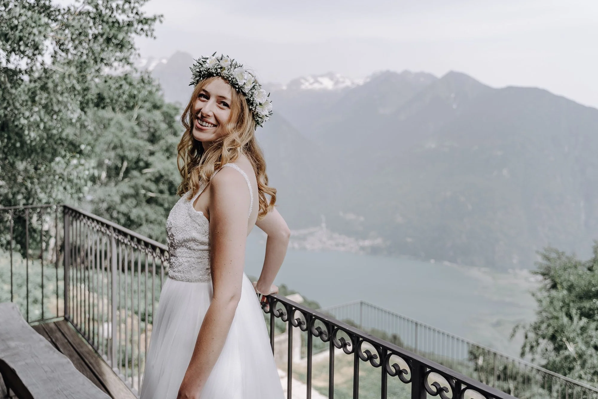 A smiling woman with long, curly blonde hair wearing a white lace wedding dress and a floral crown made of white flowers, standing on a balcony with a mountain and lake view in the background.