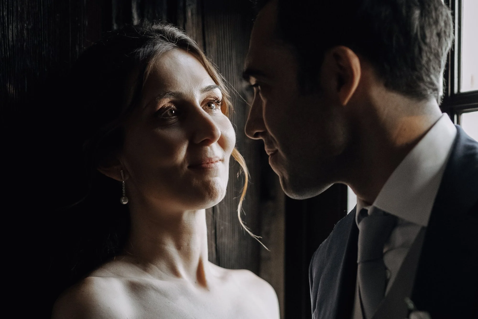 A couple sharing an intimate moment, standing close to each other with the man's forehead against the woman's face, near a window with natural light.