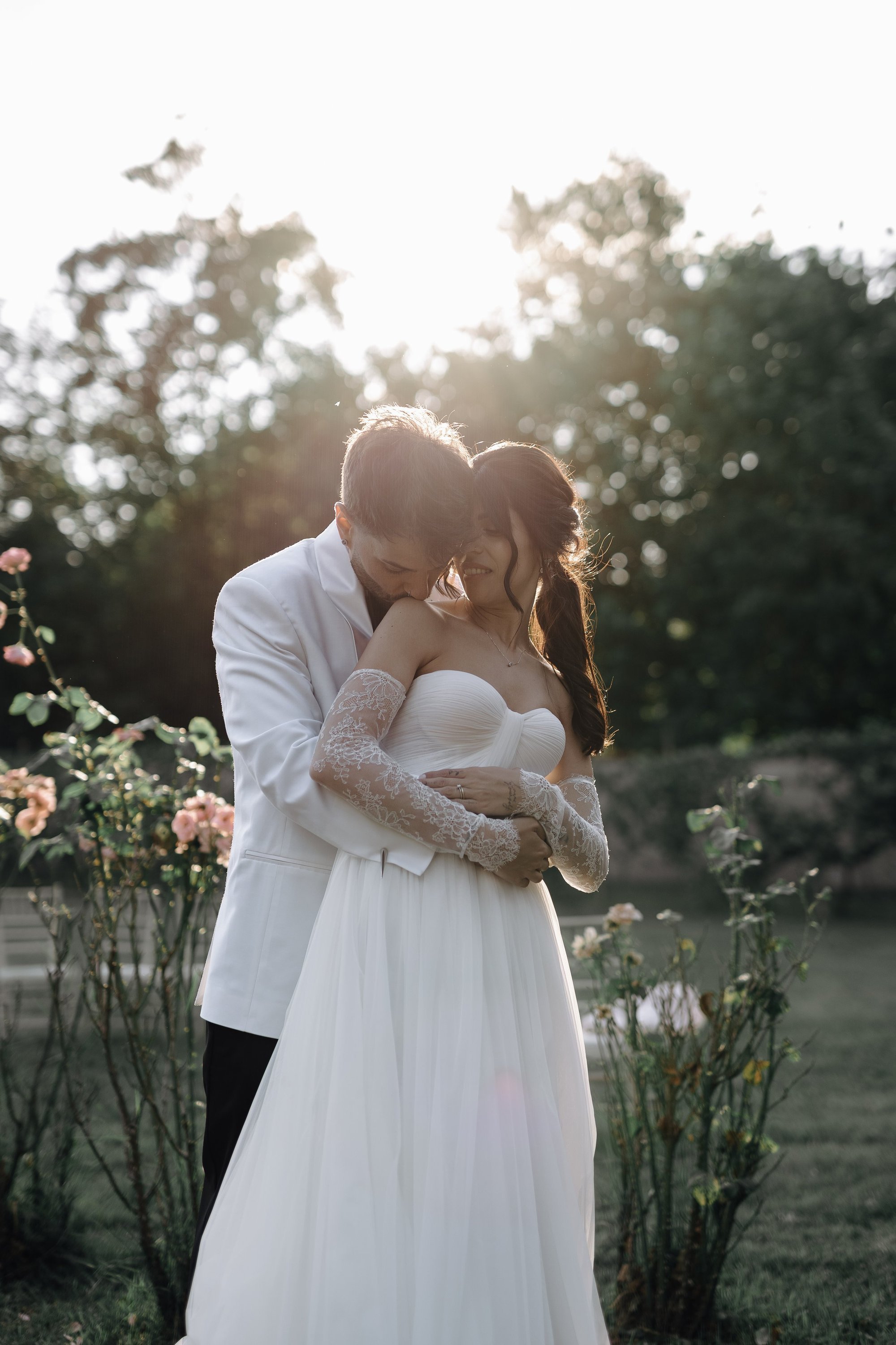 A couple dressed in wedding attire embracing outdoors during sunset, with trees and pink roses in the background.