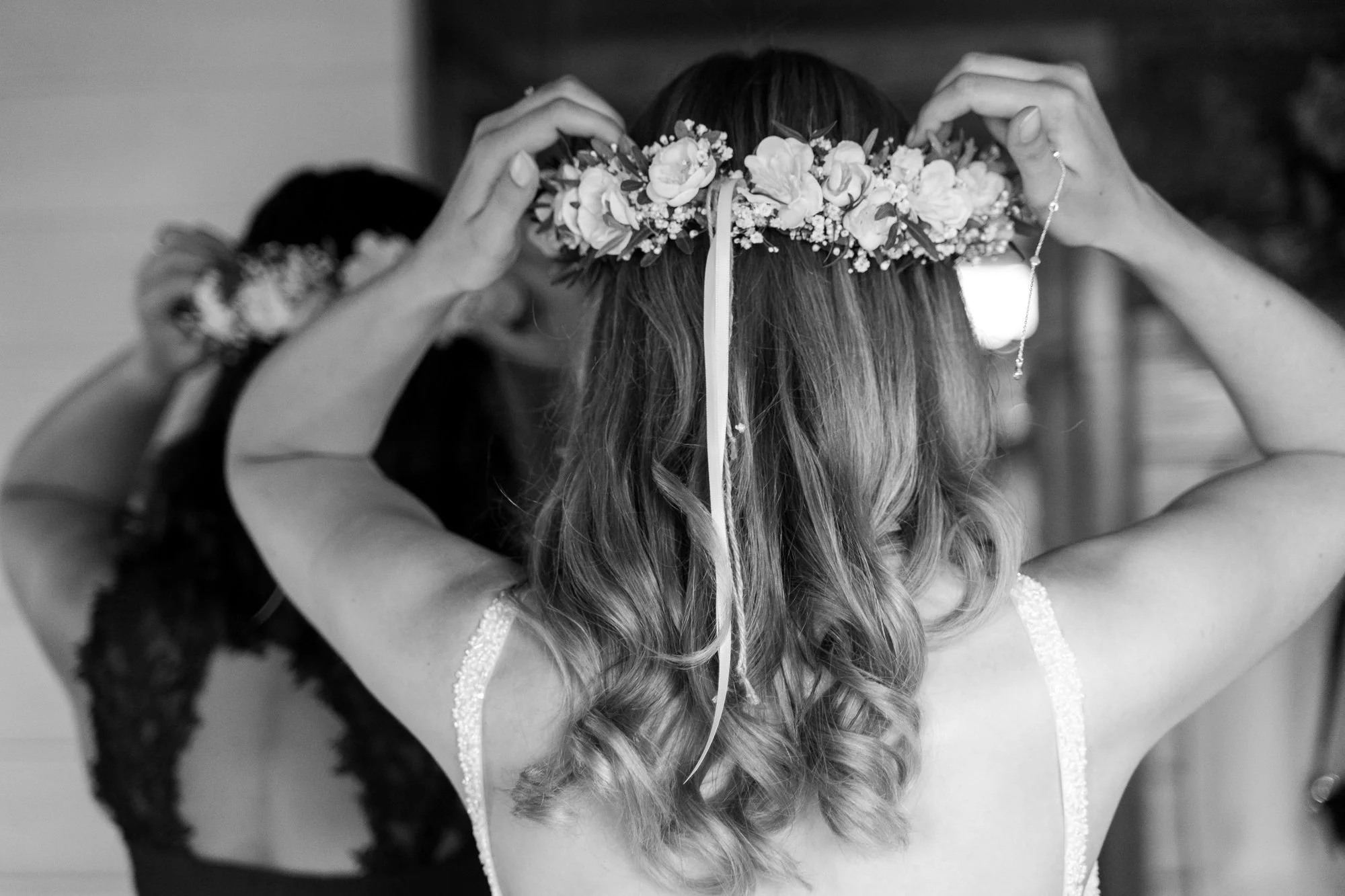 A woman wearing a floral crown is adjusting it, with two other women assisting her, seen from behind in a black and white photo.