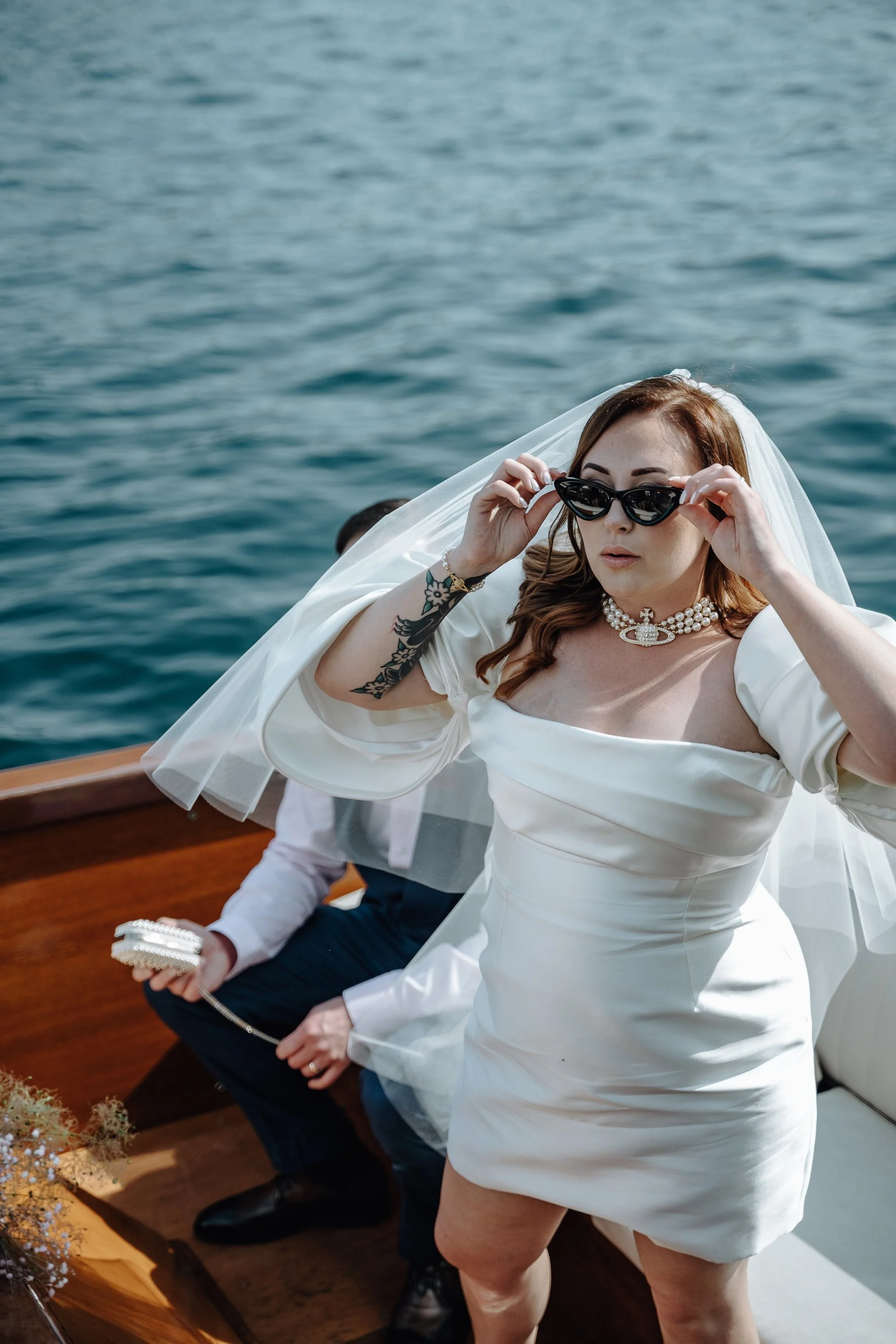 A woman in a white wedding dress and pearl necklace looking over sunglasses on a boat with water in the background.