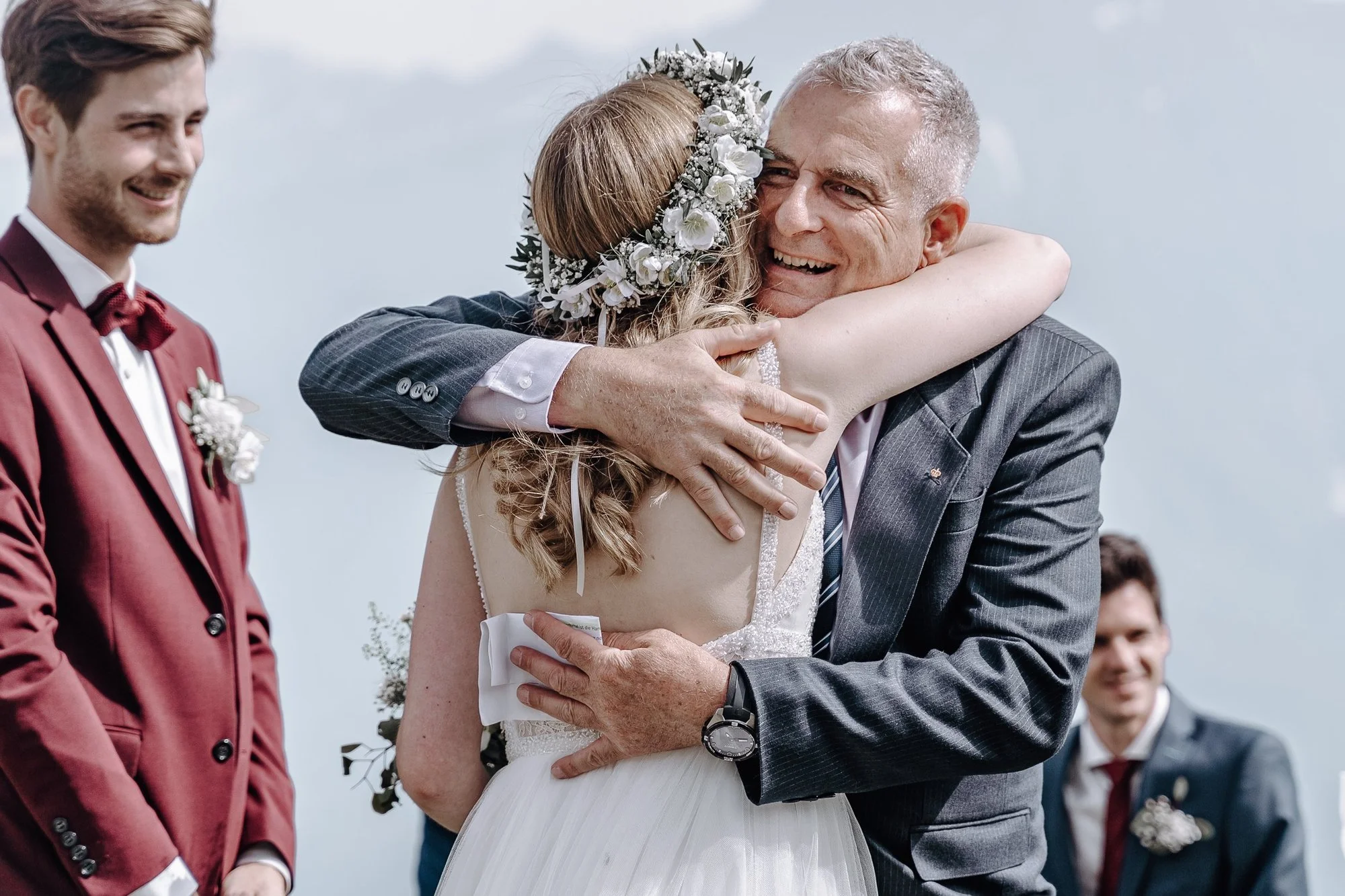 A bride and an older man, likely her father, hugging during a wedding ceremony outdoors. The bride wears a floral crown and a white gown. The groom stands to the left, wearing a burgundy suit with a boutonniere, smiling.