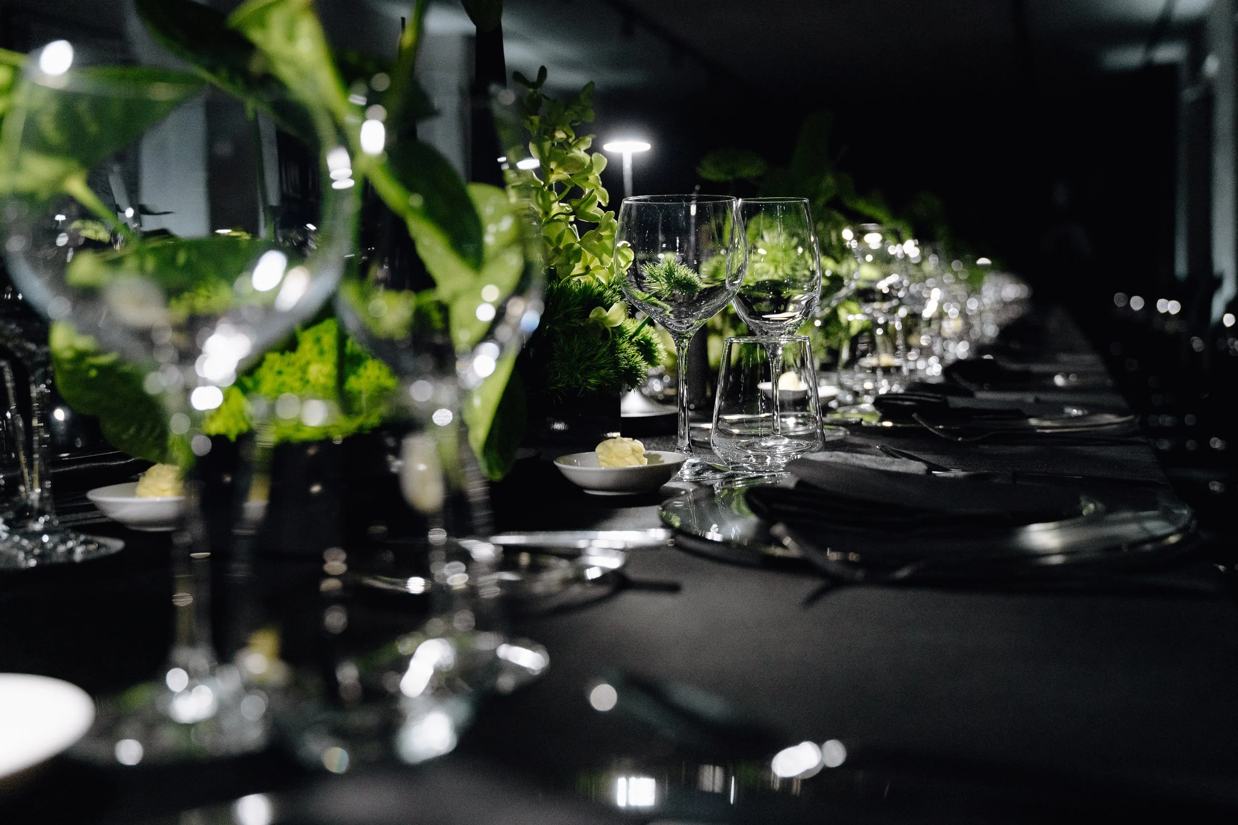 A dark table setting with glassware, greenery, and decorative lighting for a sophisticated event.