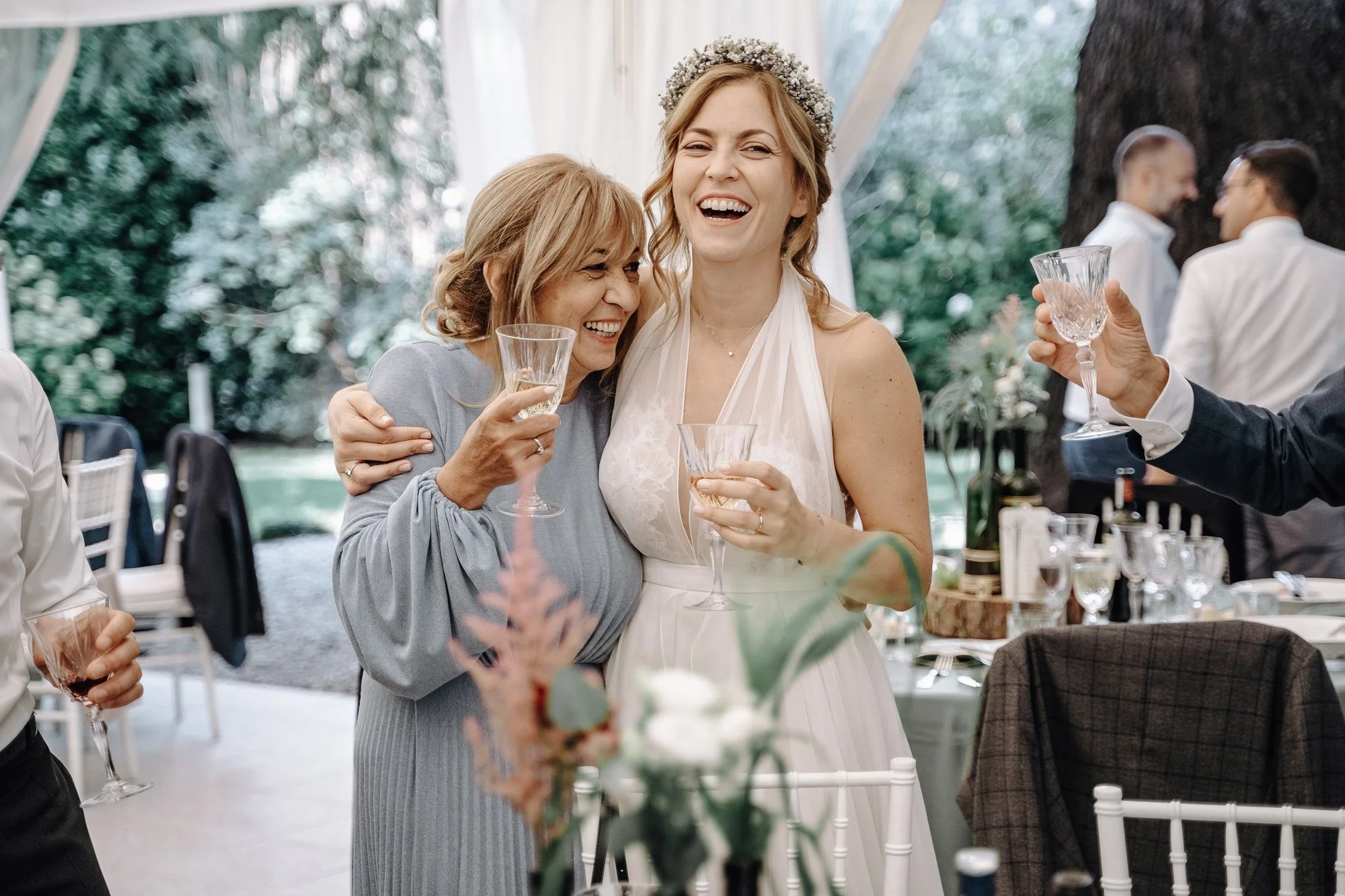 Two women at a wedding reception smiling and holding glasses of champagne. One woman is wearing a light grey dress and the other a white dress with a floral headpiece. They are embracing each other. There are other guests in the background celebratin