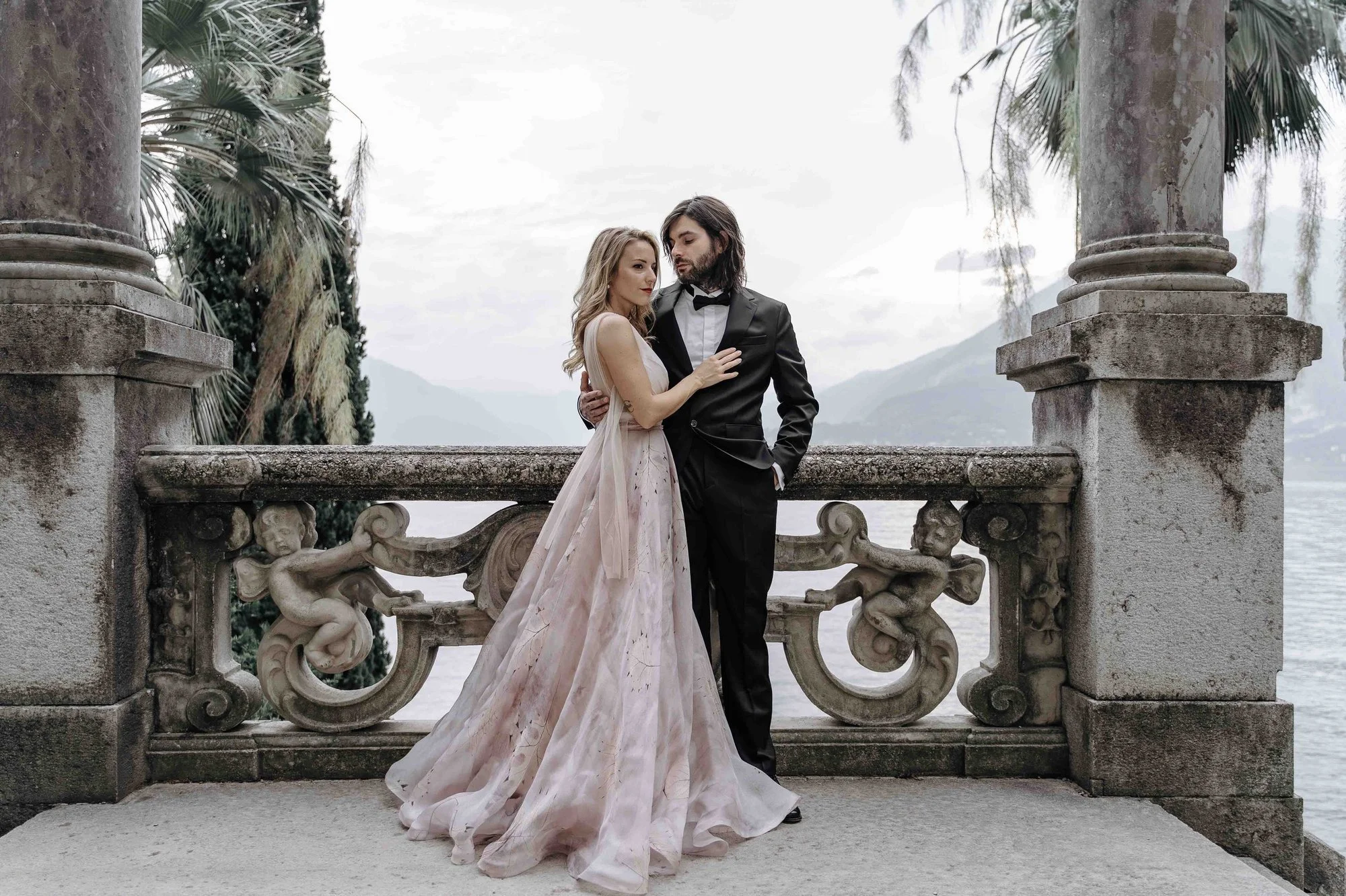 A couple in formal attire, a woman in a flowing gown and a man in a tuxedo, standing close together on a stone balcony with ornate carvings and cherub statues, overlooking a body of water and mountains in the background.