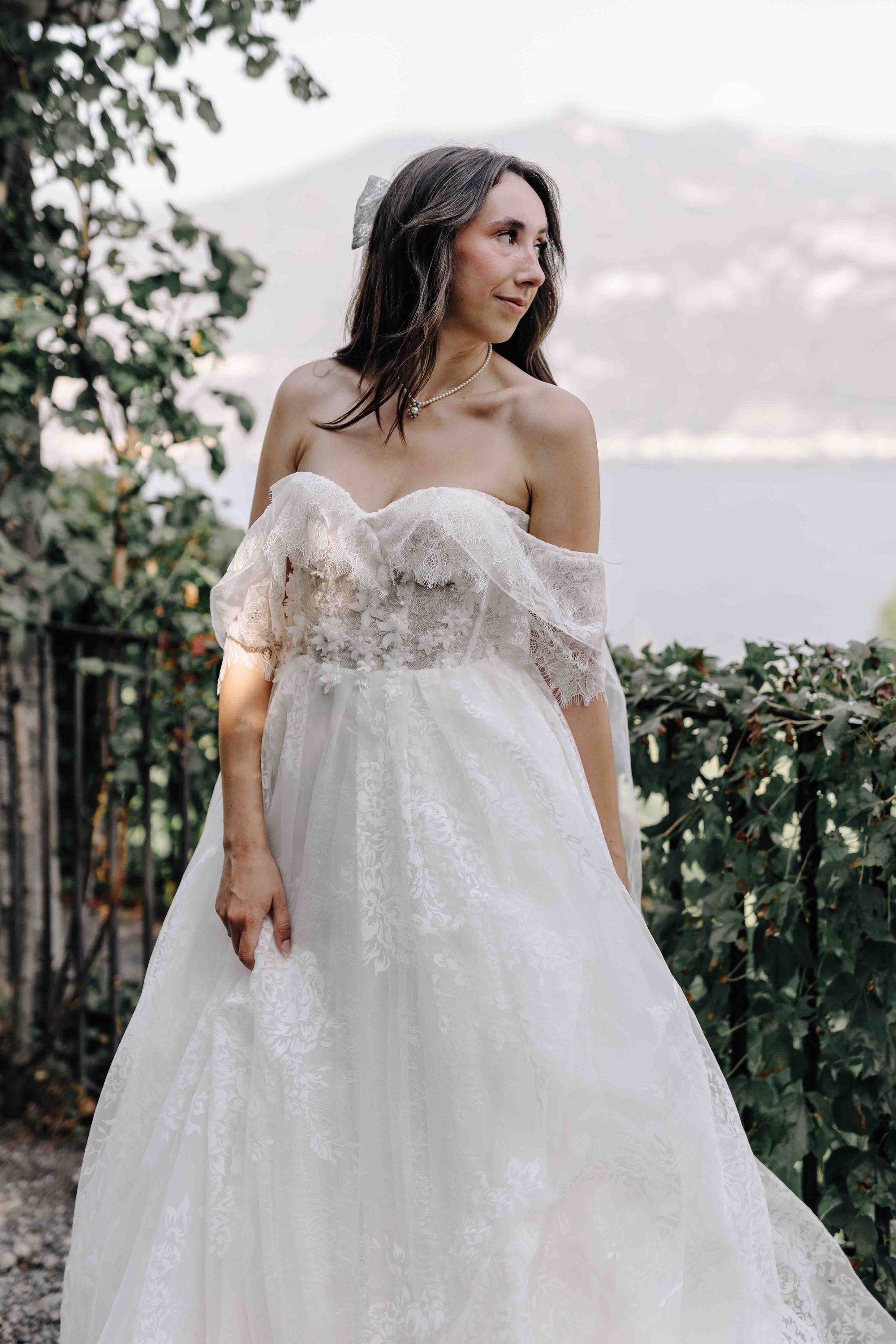 A woman in a white wedding dress stands outdoors with greenery and a body of water in the background. She has dark brown hair, a pearl necklace, and a silver hair accessory, and looks to her right with a slight smile.