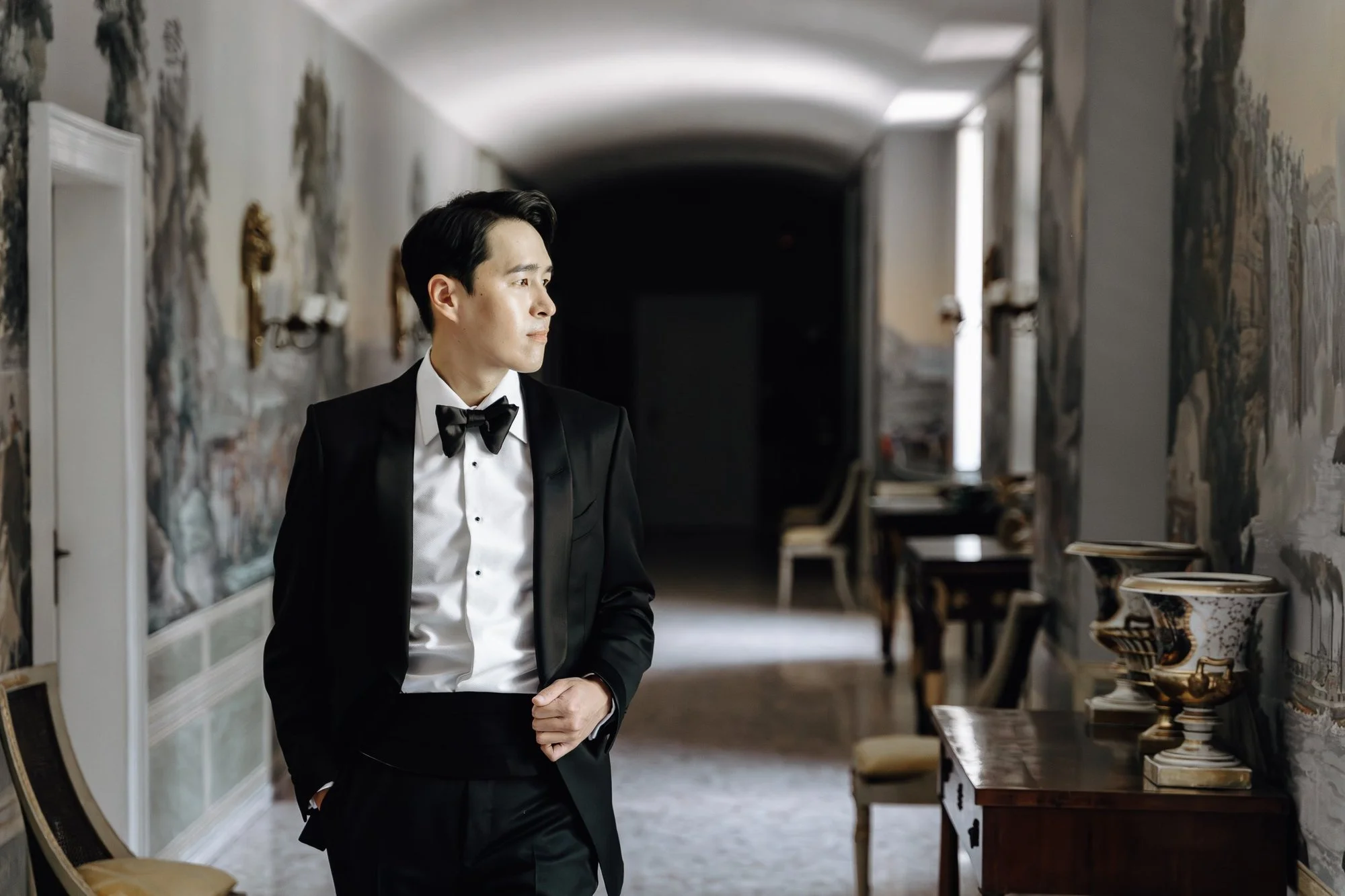 A young man dressed in a tuxedo standing in an art gallery or museum, surrounded by decorative items and paintings on the walls.